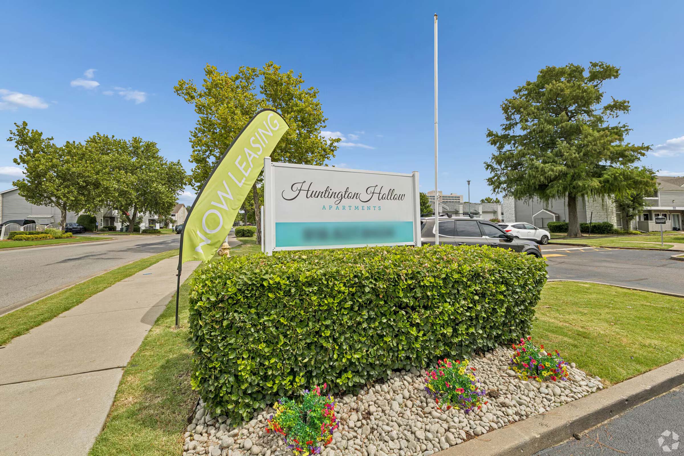 Sign for Huntington Hollow Apartments with a "Now Leasing" banner. The sign is surrounded by well-kept shrubs and a landscaped area. Clear blue sky and a few clouds are visible in the background, indicating a sunny day.