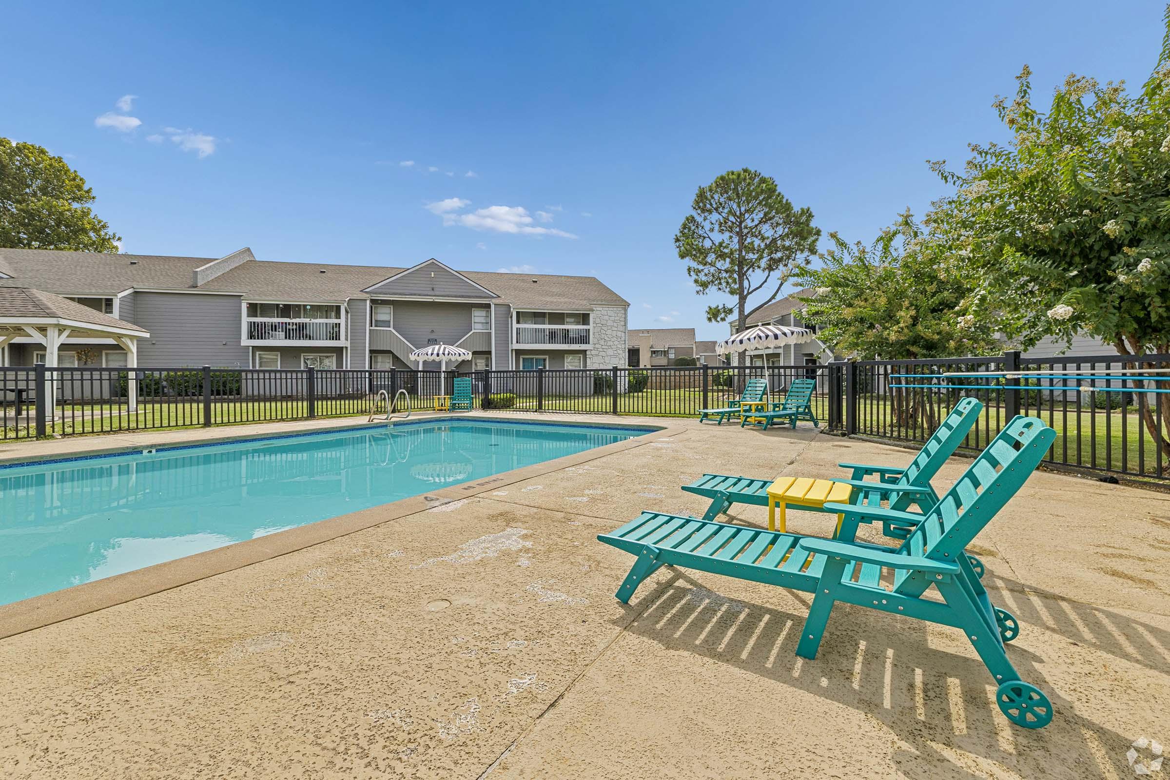 A tranquil pool area featuring a well-maintained swimming pool surrounded by green grass. Bright turquoise lounge chairs are arranged around the pool, and there are umbrellas for shade. In the background, there are residential buildings and trees under a clear blue sky.