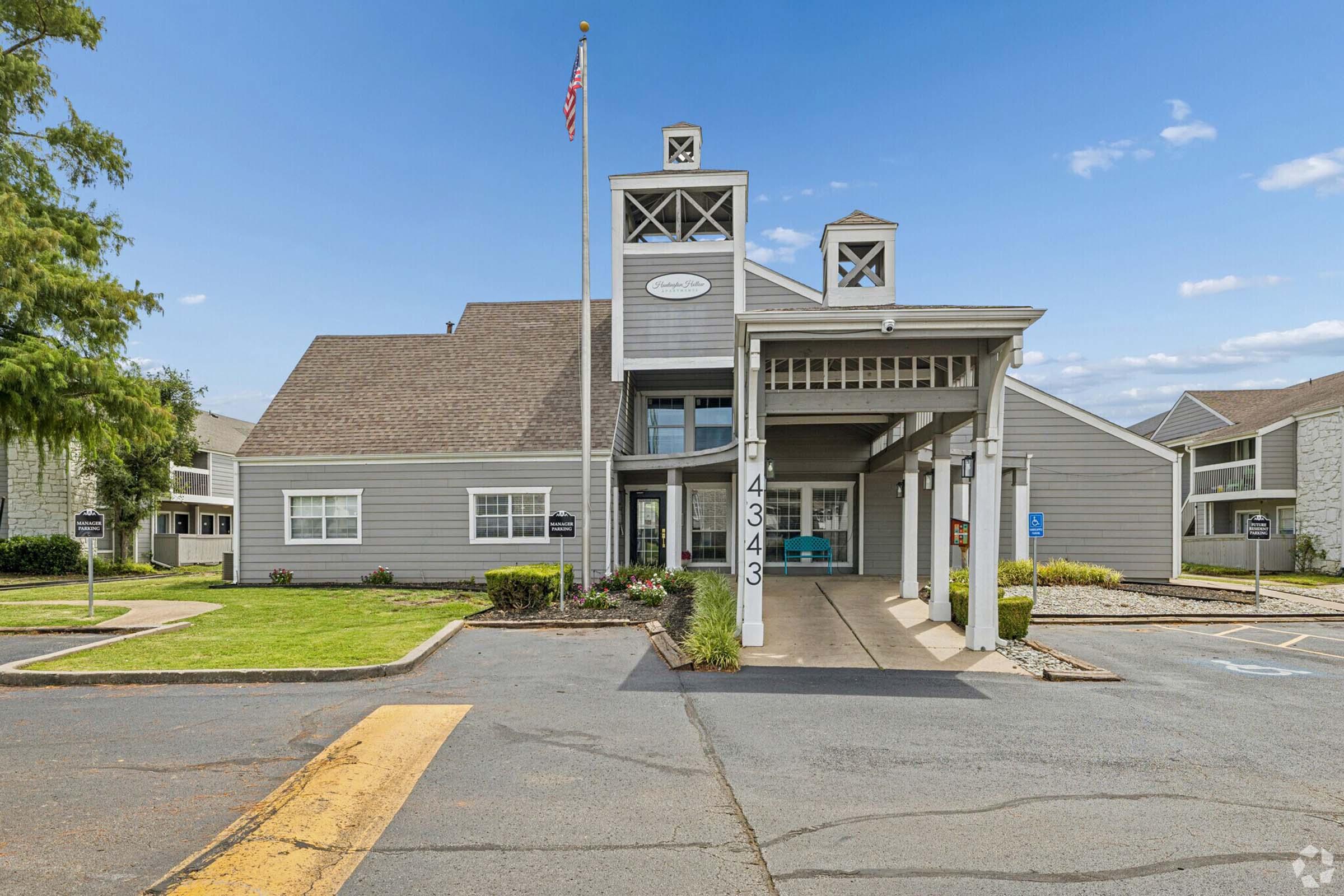 A modern apartment complex entrance featuring a gray building with a peaked roof, an American flag flying in front, and a landscaped area. The pathway leads to a covered entryway, with parking spaces visible on the side. Clear blue skies and greenery enhance the inviting atmosphere of the property.