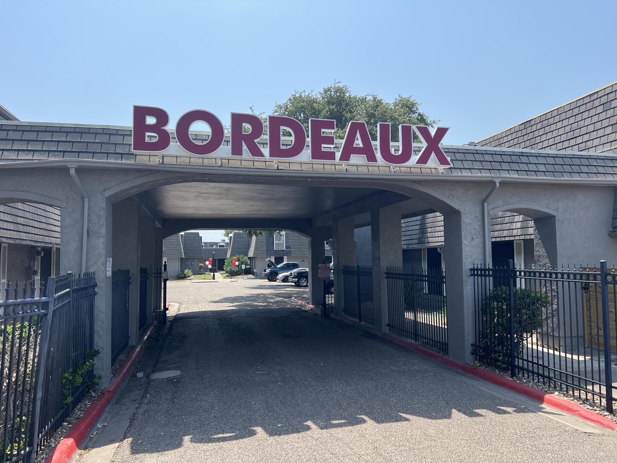 Signage reading "BORDEAUX" above an entrance, leading into a parking area flanked by buildings. The scene is sunny, with blue skies and some greenery. Several cars are parked in the lot, and there is a fence along the walkway.