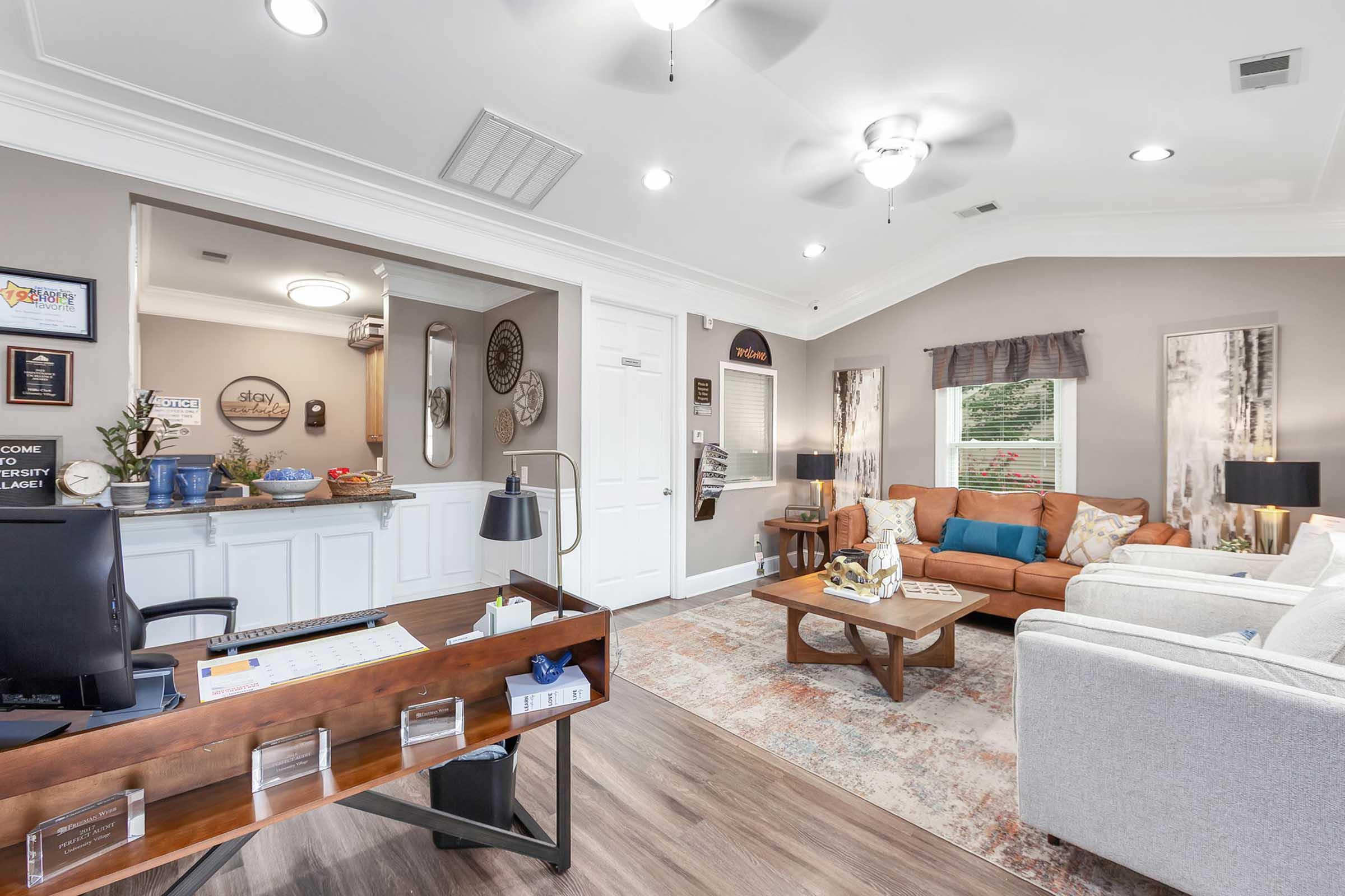 A cozy office space featuring a wooden desk with a lamp, two comfortable sofas, and a coffee table. The walls are painted in a soft gray and decorated with framed art. Natural light filters through the windows, and a small kitchen area is visible in the background, creating a welcoming atmosphere.