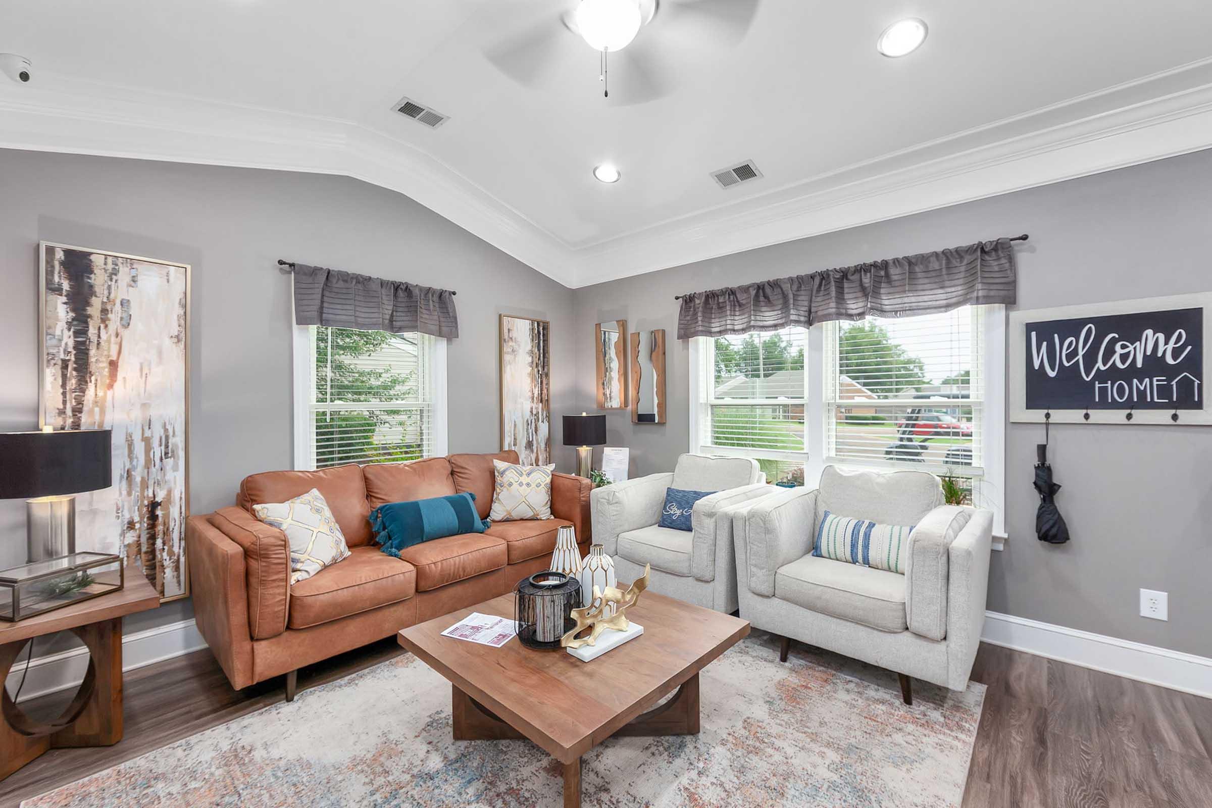 A cozy living room featuring a light gray color scheme with a brown leather sofa and two light-colored armchairs. There is a wooden coffee table with decorative items and a welcome sign on the wall. Natural light streams in through large windows, enhancing the inviting atmosphere of the space.
