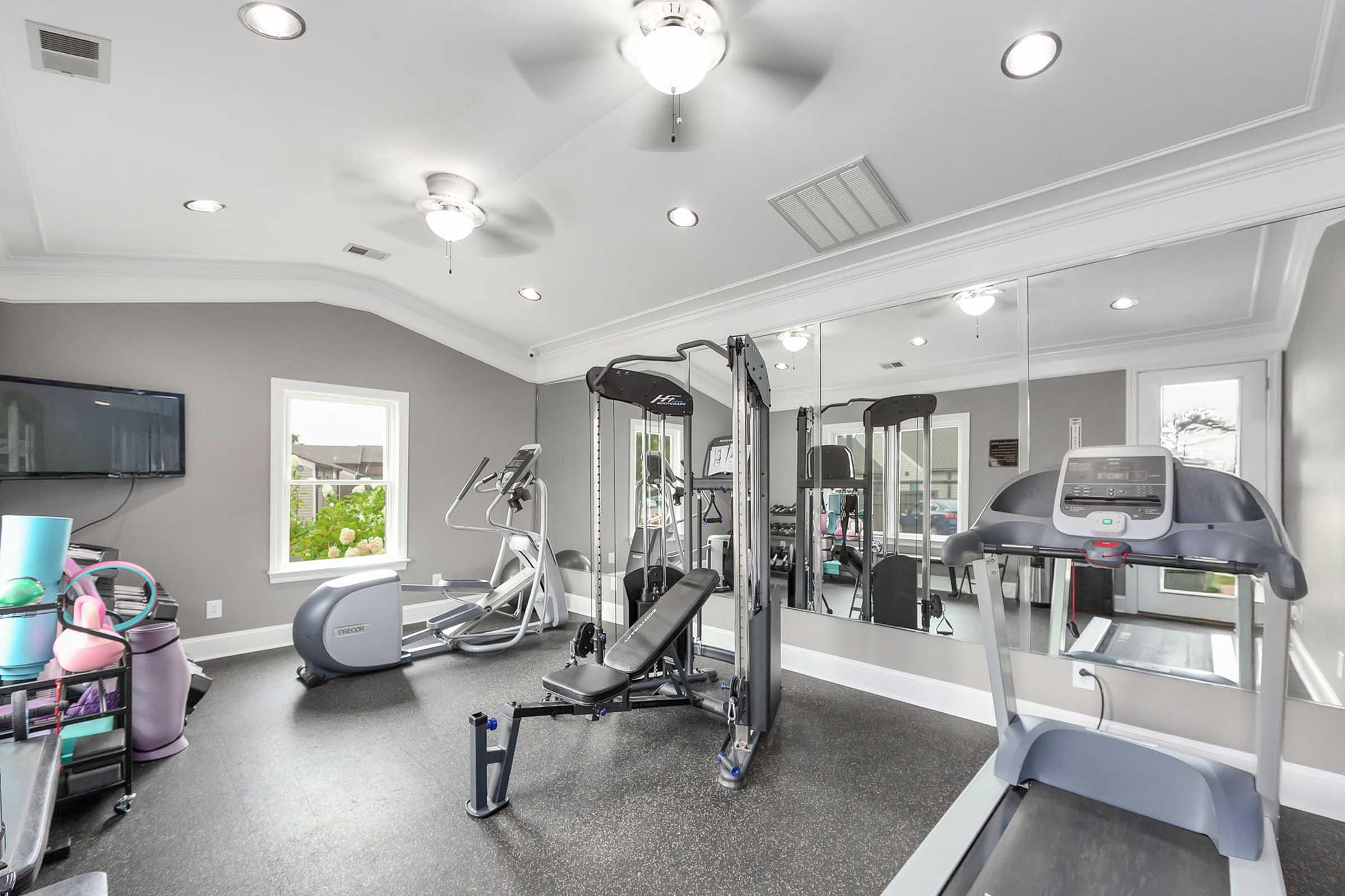 A modern home gym featuring a variety of exercise equipment, including a treadmill, elliptical machine, and multi-station weight machine. The room has a light gray color scheme, ceiling fan, and mirrors reflecting the equipment. Natural light comes in from a window, enhancing the workout space.