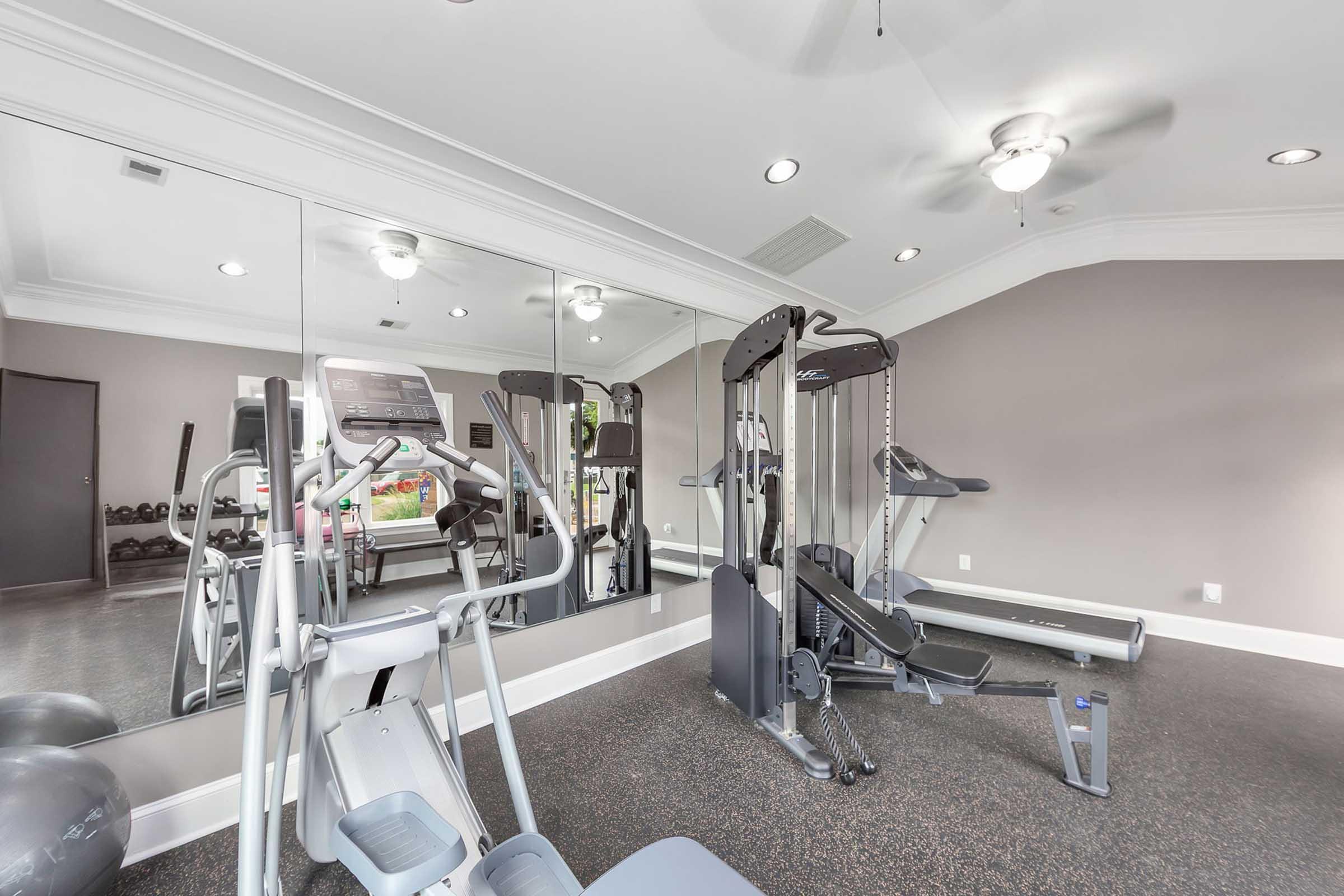A well-lit gym interior featuring exercise equipment. There are cardio machines including a treadmill and an elliptical trainer, strength training machines, and free weights. Mirrors on the wall enhance the space, and the floors are covered with dark carpet. Ceiling fans provide ventilation.
