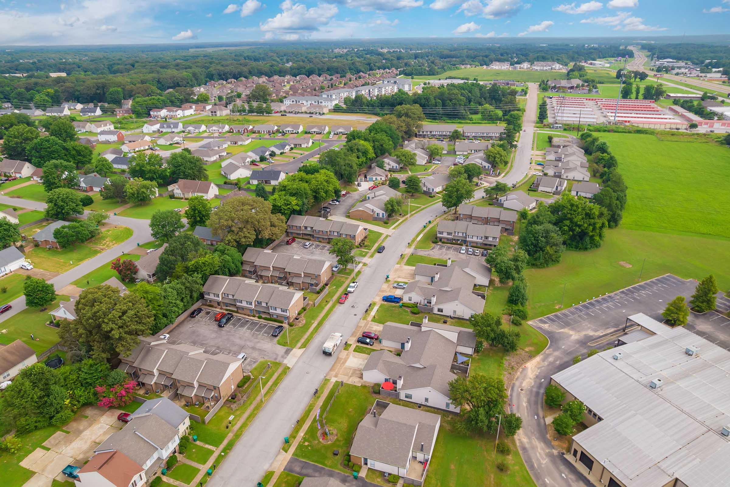 Aerial view of a suburban neighborhood featuring low-rise residential buildings, tree-lined streets, and a well-maintained landscape. In the background, a stretch of commercial development and open green fields can be seen, with a mix of homes and parking areas throughout the area. Bright, partly cloudy sky above.
