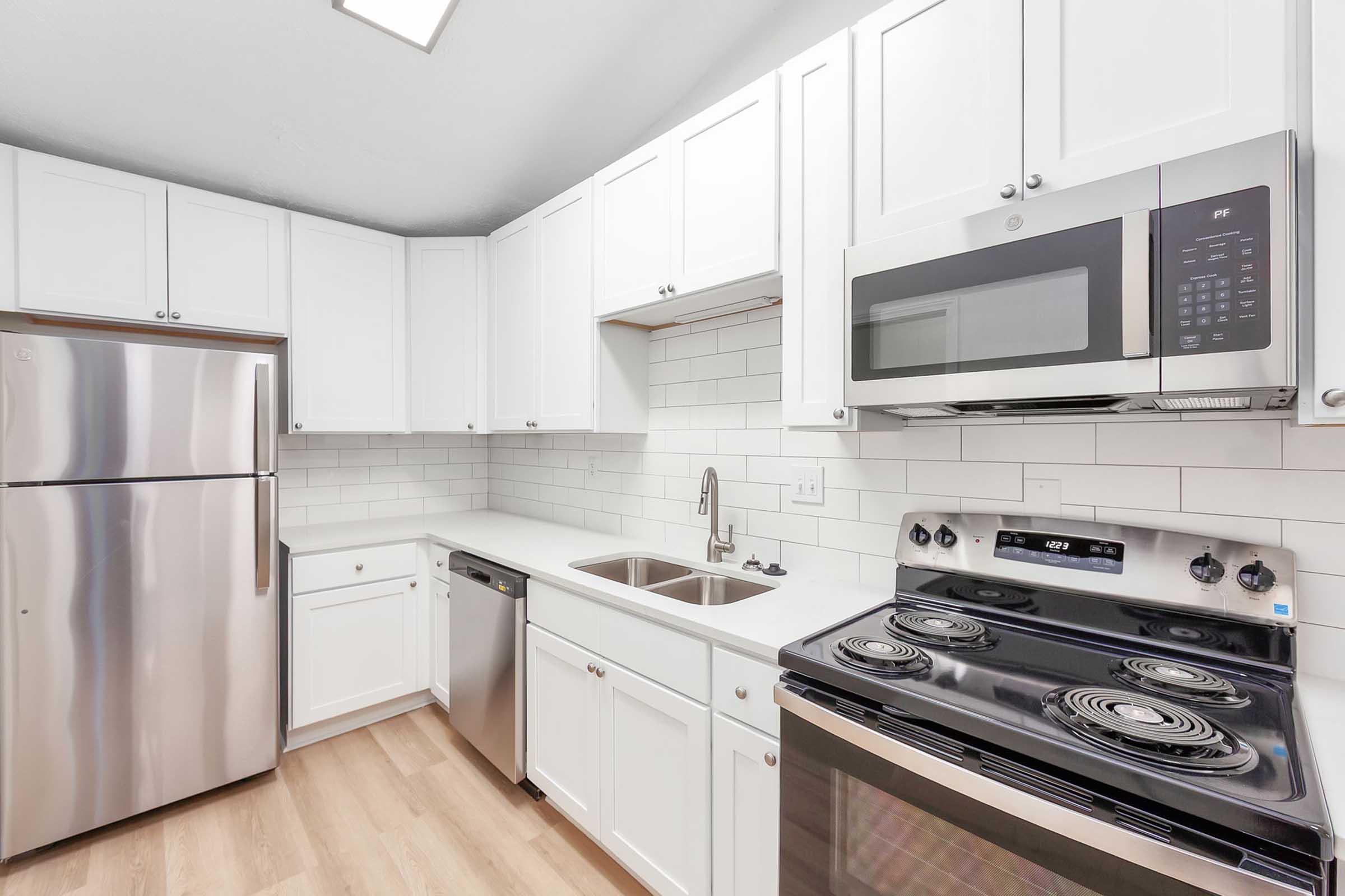 Modern kitchen featuring white cabinets, stainless steel appliances including a refrigerator, microwave, and stove. A double sink is integrated into the countertop, with a minimalist tile backsplash and wooden flooring, creating a bright and functional space.