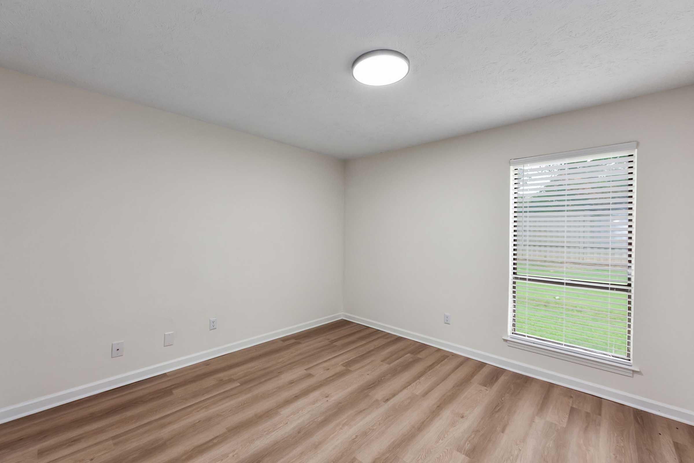 Bright and empty room with light-colored walls, wooden flooring, and a large window with blinds allowing natural light. A flush ceiling light is installed, enhancing the illumination in the space. The room appears ready for furnishing and decoration.