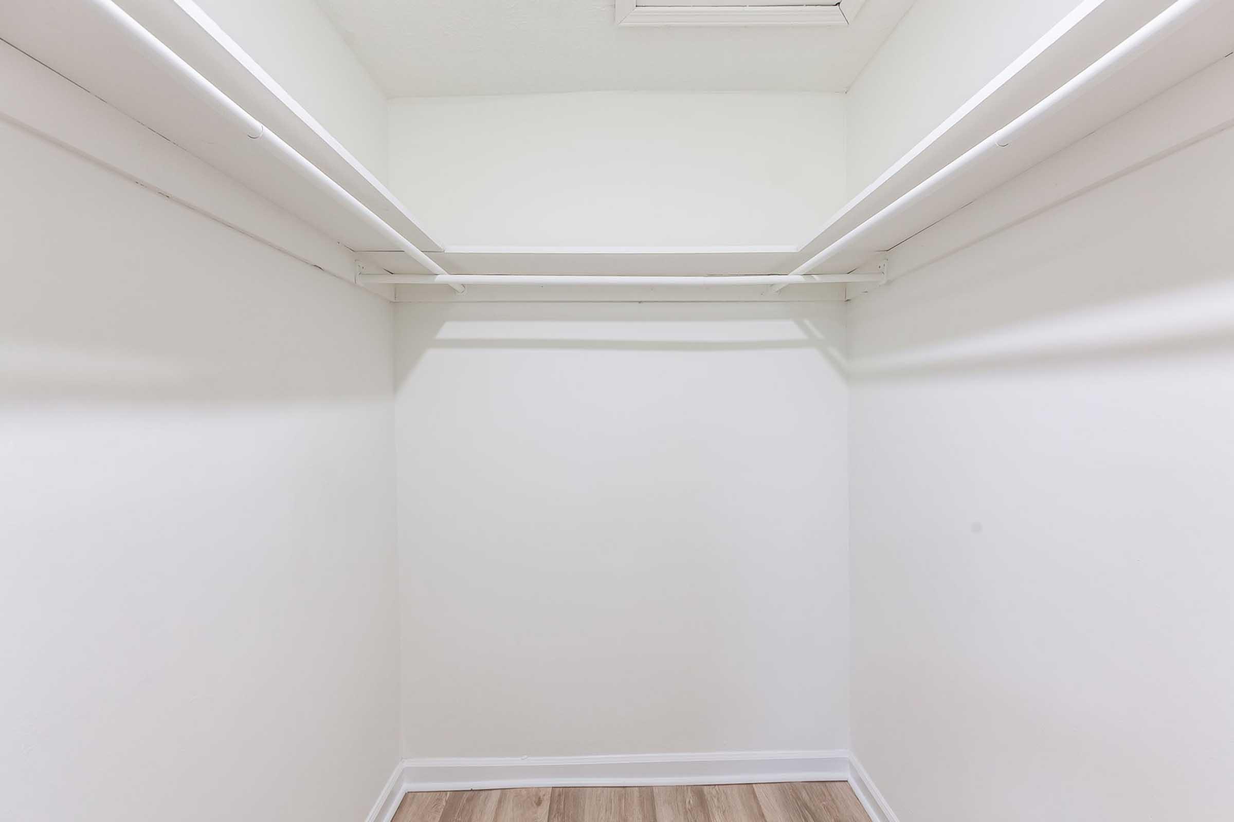 Empty closet with white walls and two hanging shelves. The floor features wood-like laminate, and there is a small ceiling vent visible. The space appears clean and minimally furnished.