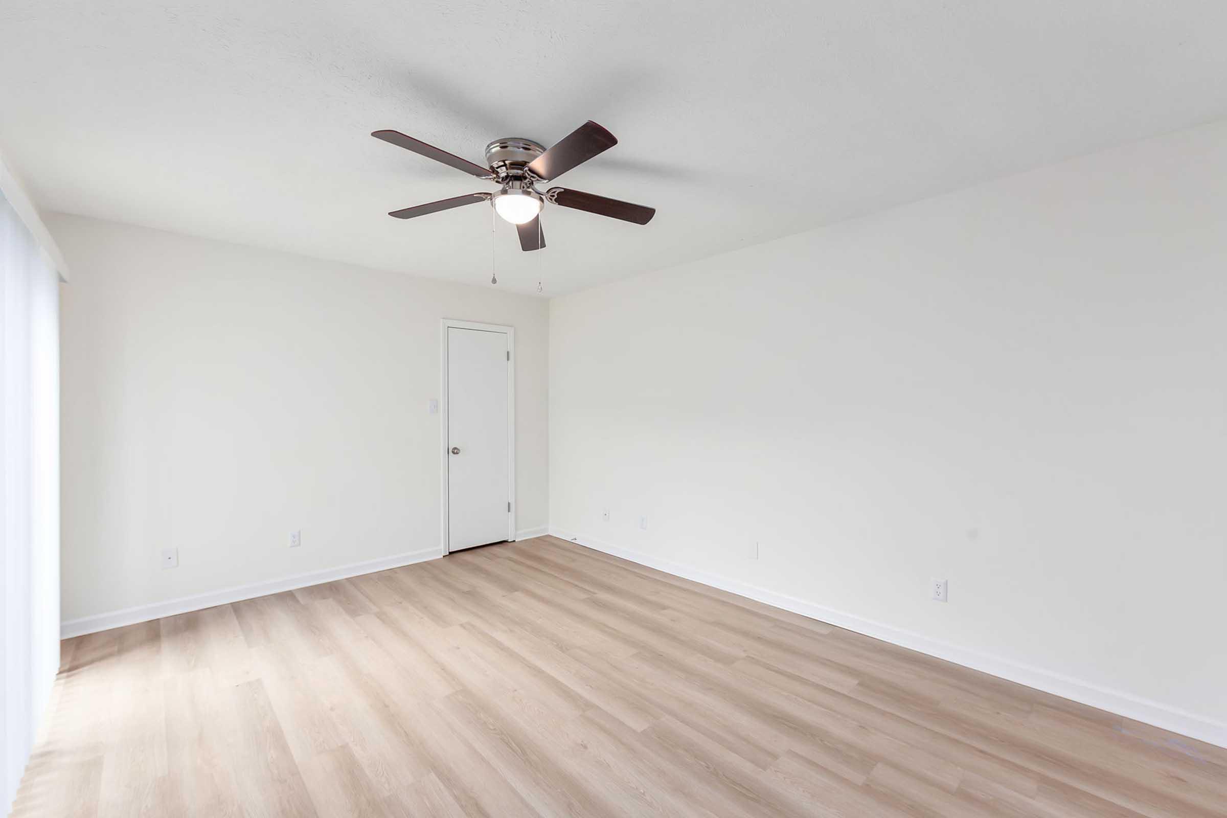 Empty room with light wood flooring, a ceiling fan with five blades, and a white wall. A door is partially visible on the right side. Natural light comes through a window on the left, illuminating the space. The overall atmosphere is minimalistic and bright.