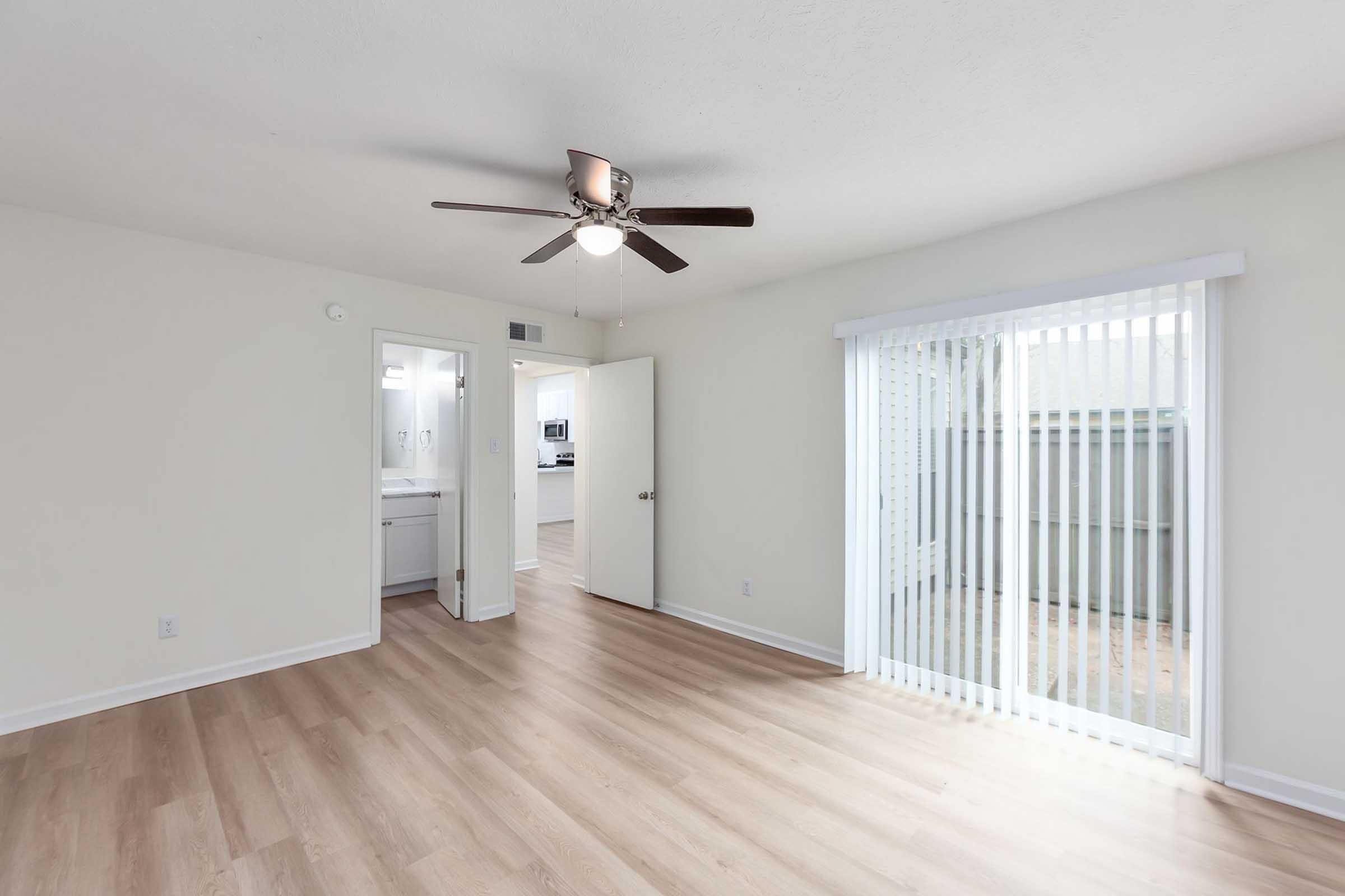 A well-lit, empty living room featuring light wood flooring, a ceiling fan, and large sliding glass doors leading outside. A doorway on the left leads to a bathroom or kitchen area, with modern fixtures visible. The walls are painted a soft white, creating a bright and open atmosphere.