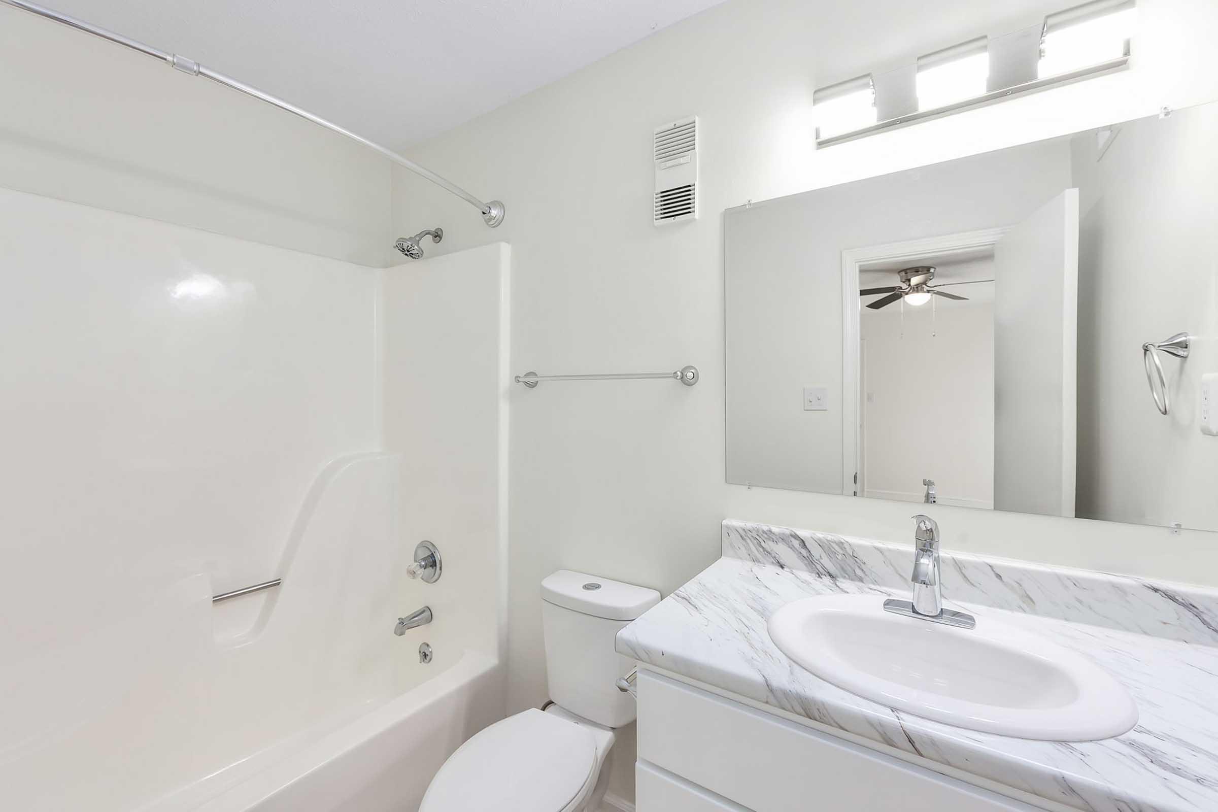 A modern bathroom featuring a white bathtub with a shower, a marble countertop sink, a toilet, and a mirror above the sink. The walls are painted light colors, and there is a ceiling fan visible through an open door. The overall appearance is clean and well-lit, with a minimalist design.