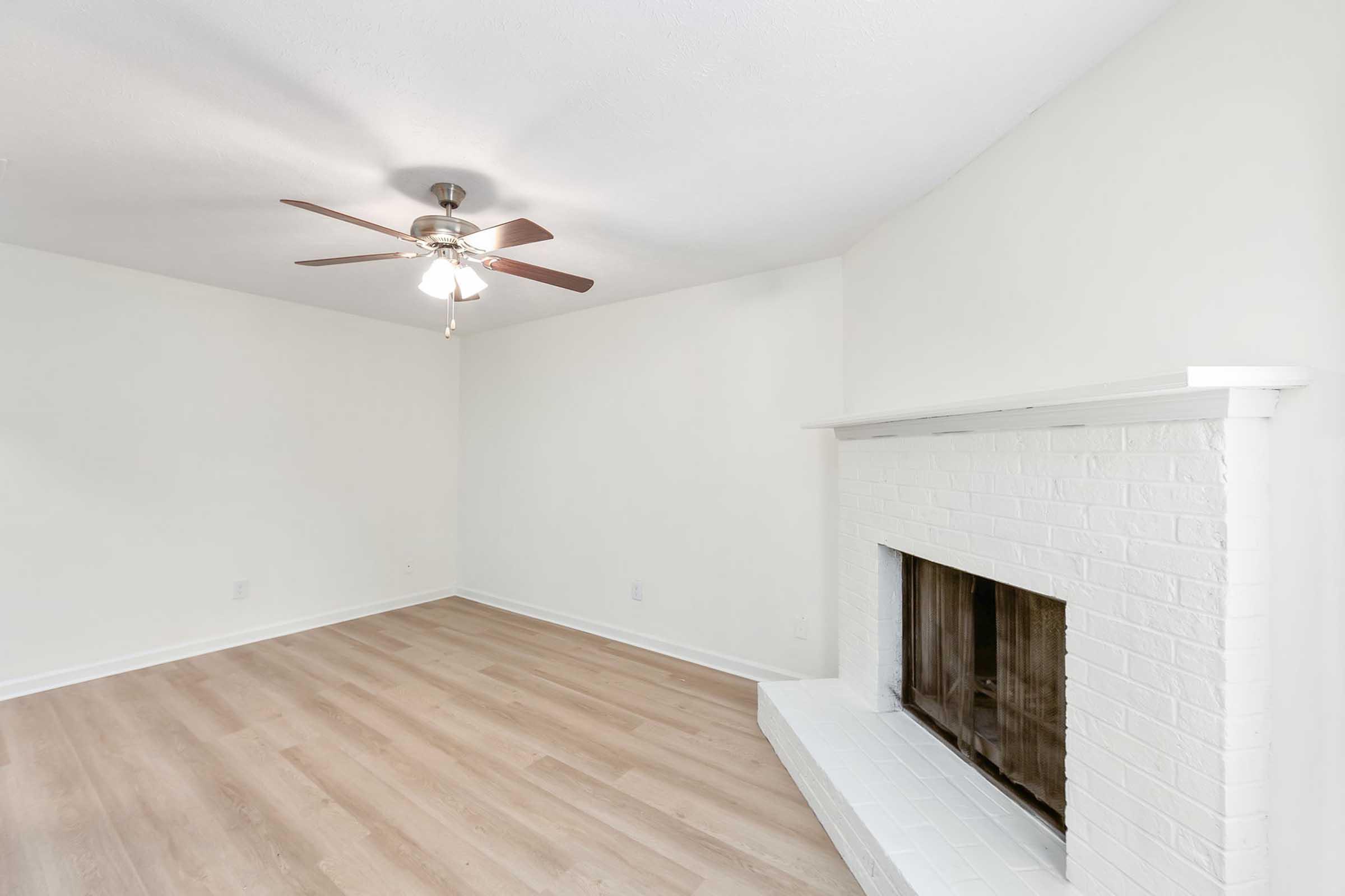 Bright, spacious living room with light wood flooring, white walls, and a ceiling fan. A white brick fireplace serves as a focal point on one wall, enhancing the cozy atmosphere. The room is uncluttered, providing a blank canvas for potential furnishings and decor.