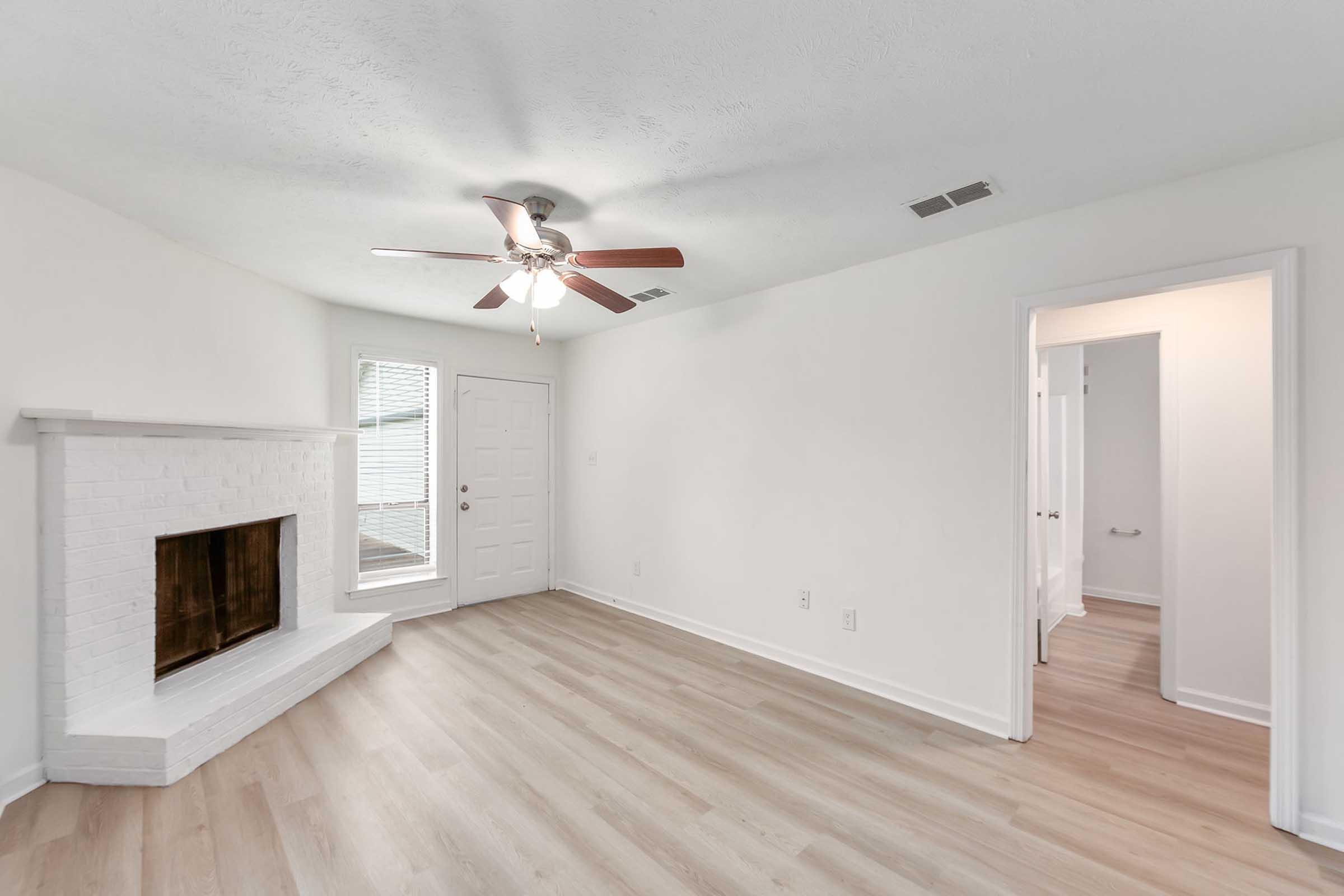A bright, empty living room with light-colored walls and new hardwood flooring. Features include a white brick fireplace, a ceiling fan, and a door leading outside. A doorway on the right opens to another room or hallway. Natural light enters through a nearby window.