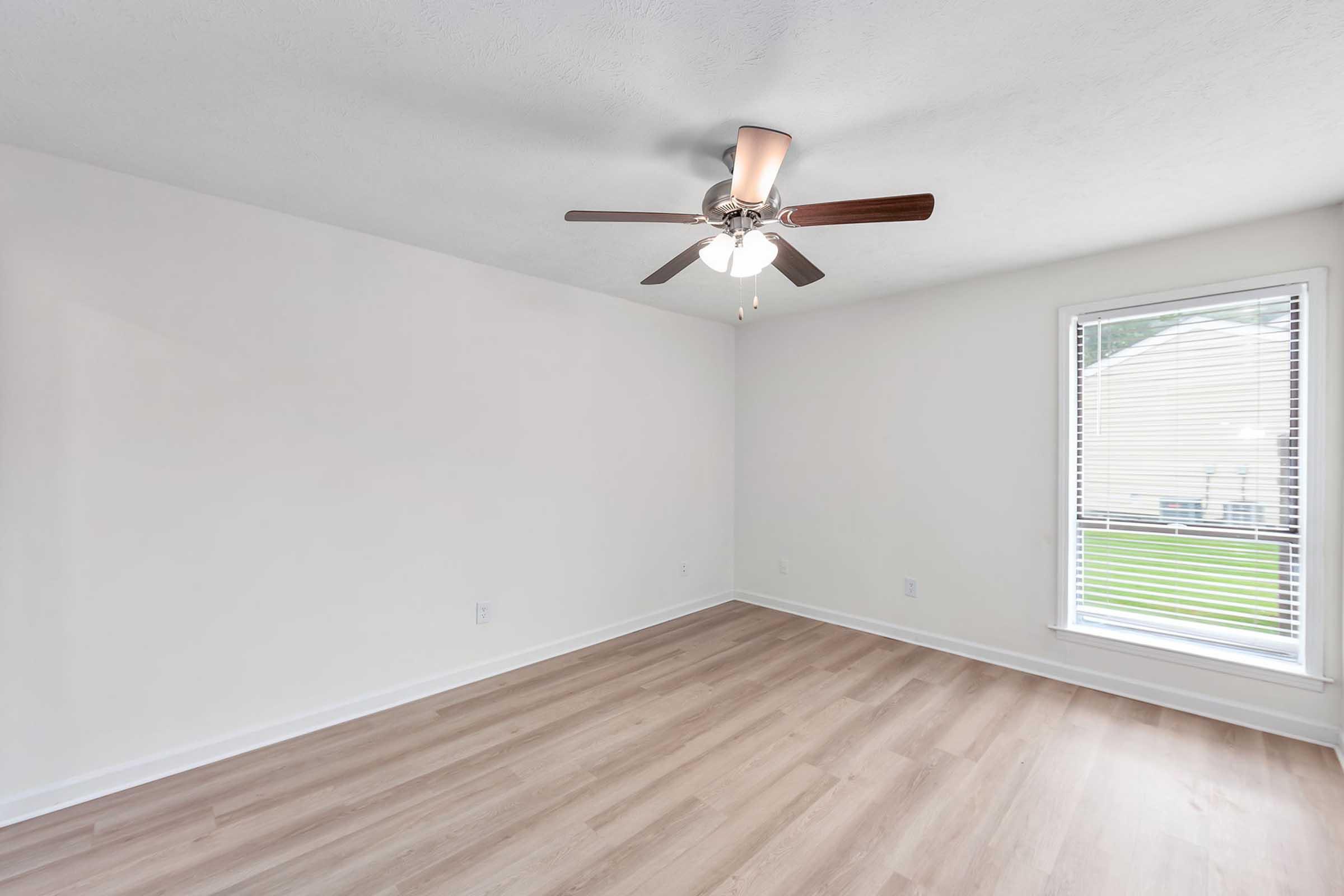 A spacious, empty room with light wooden flooring and white walls. There is a ceiling fan with five blades, and a window with blinds allows natural light to illuminate the space. The room has a simple and modern design, creating a bright and airy atmosphere.