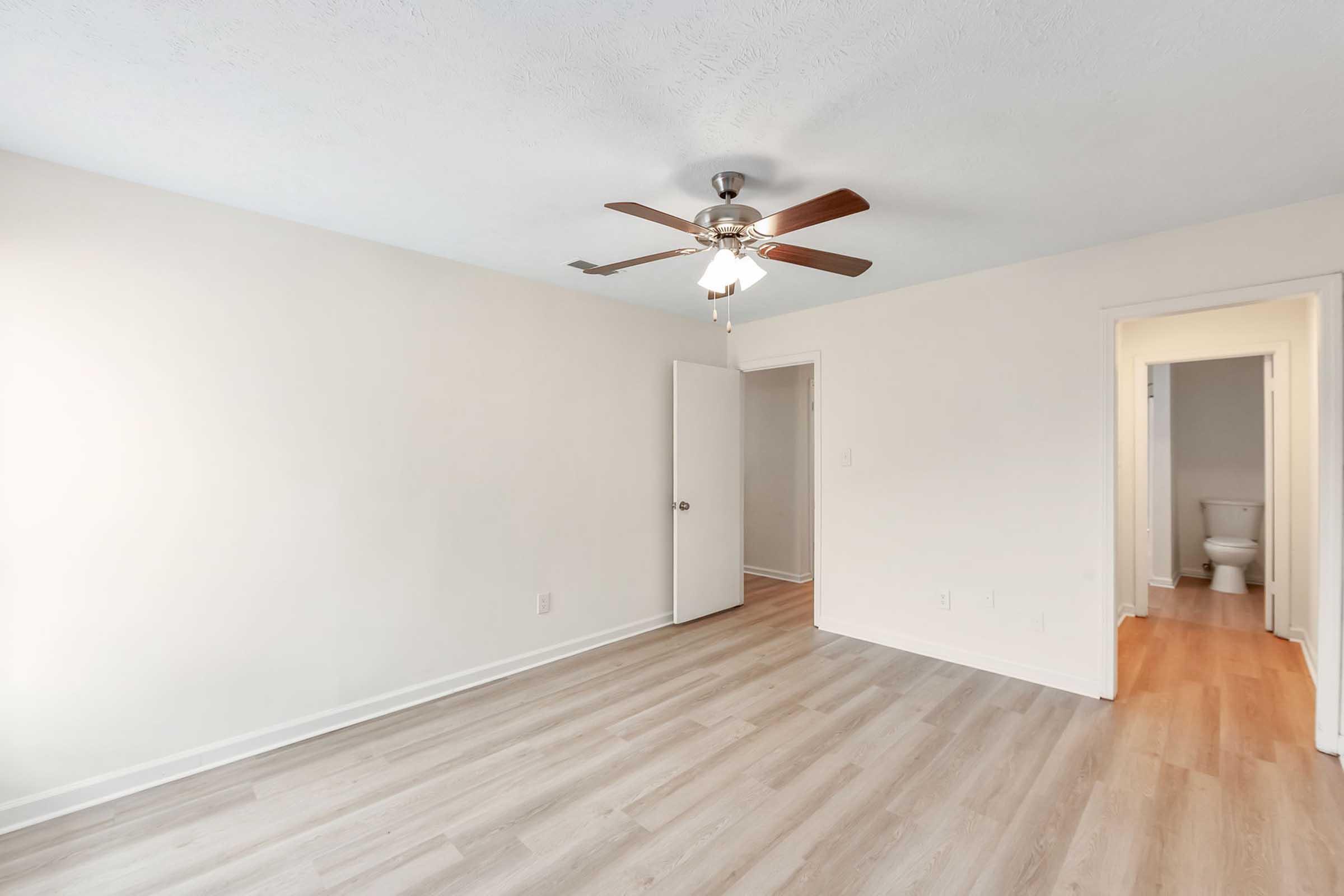 A spacious, well-lit room with light-colored walls and a ceiling fan. The flooring is a mix of wood and laminate. A closed door leads to another room, while an open door in the background reveals a bathroom. The overall atmosphere is bright and inviting, ideal for various interior design options.