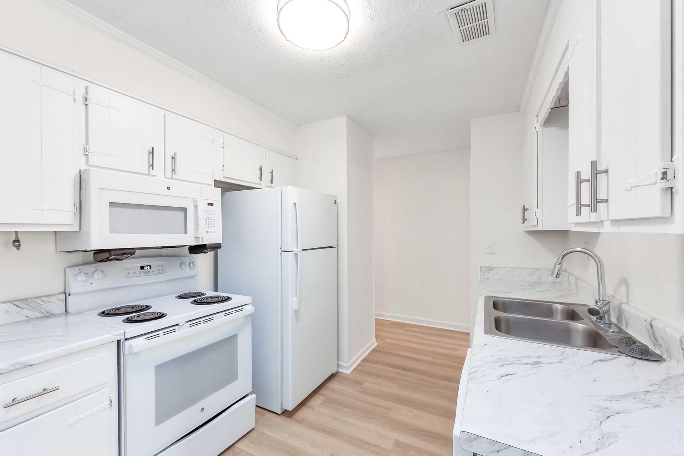 A modern kitchen featuring white cabinets, a white stove and microwave, and a refrigerator. The countertop has a marble finish, and there are dual sinks. The room has light-colored flooring and is well-lit with a ceiling fixture.