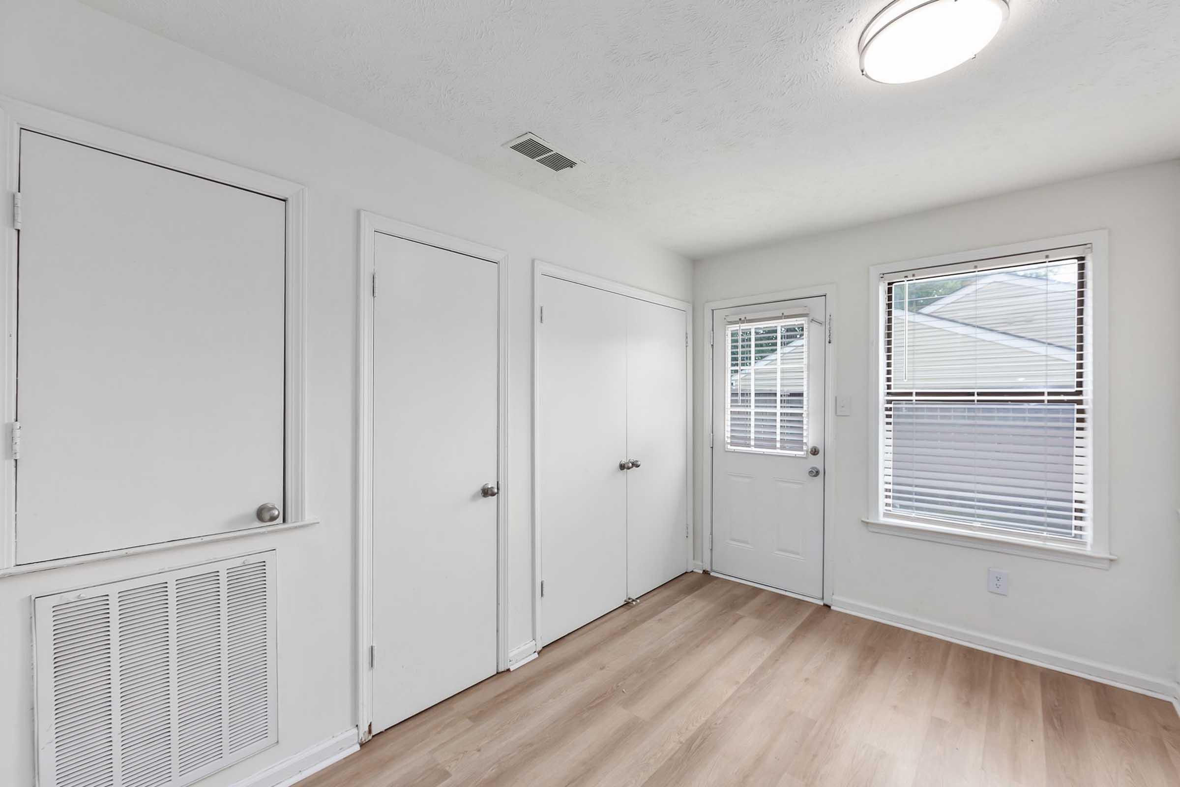 Interior of a room featuring beige walls and light wooden flooring. There are three closed doors on one side and a door leading outside on the other. A ceiling light provides illumination, and a vent is visible on the wall. Natural light enters through the window near the door.