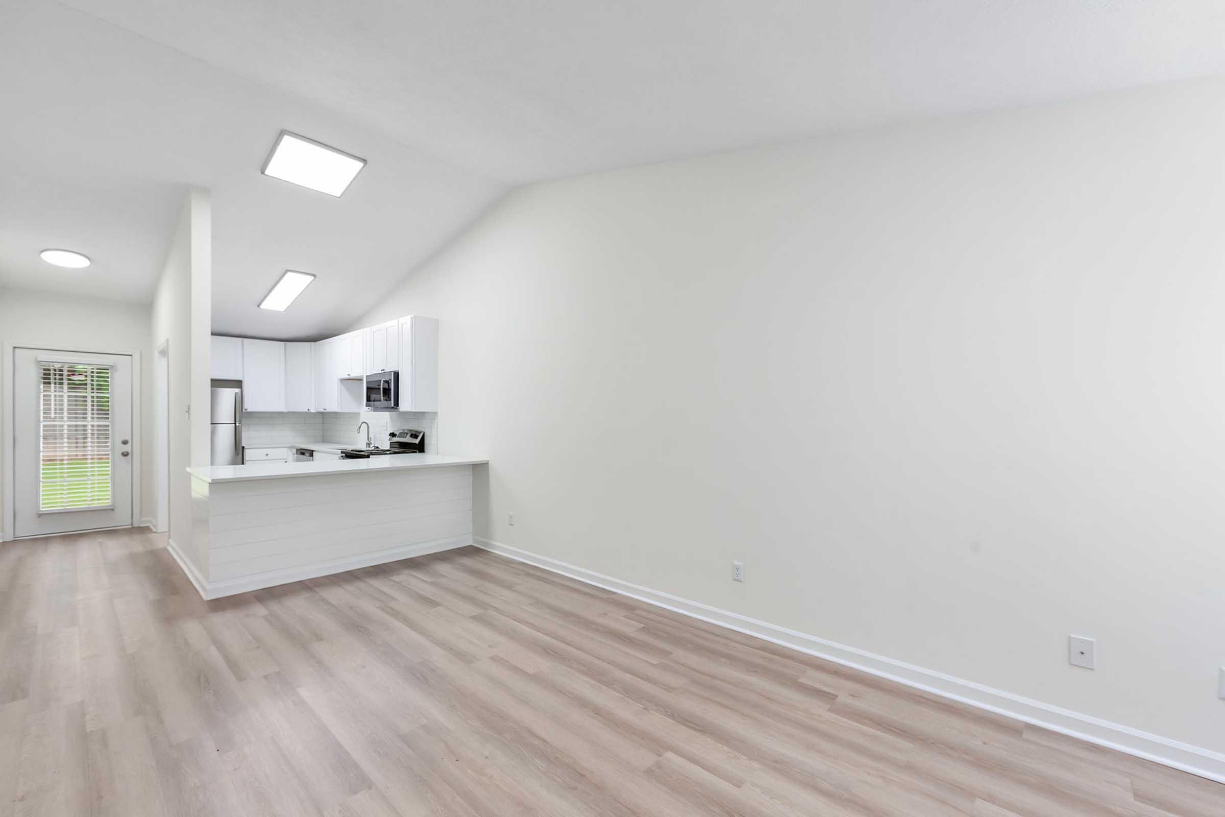 Bright and spacious interior of a modern living area featuring light-colored walls, hardwood flooring, and a sleek kitchen with white cabinets. The scene includes an open layout, large windows, and a door providing access to an outdoor space. Natural light enhances the airy atmosphere.