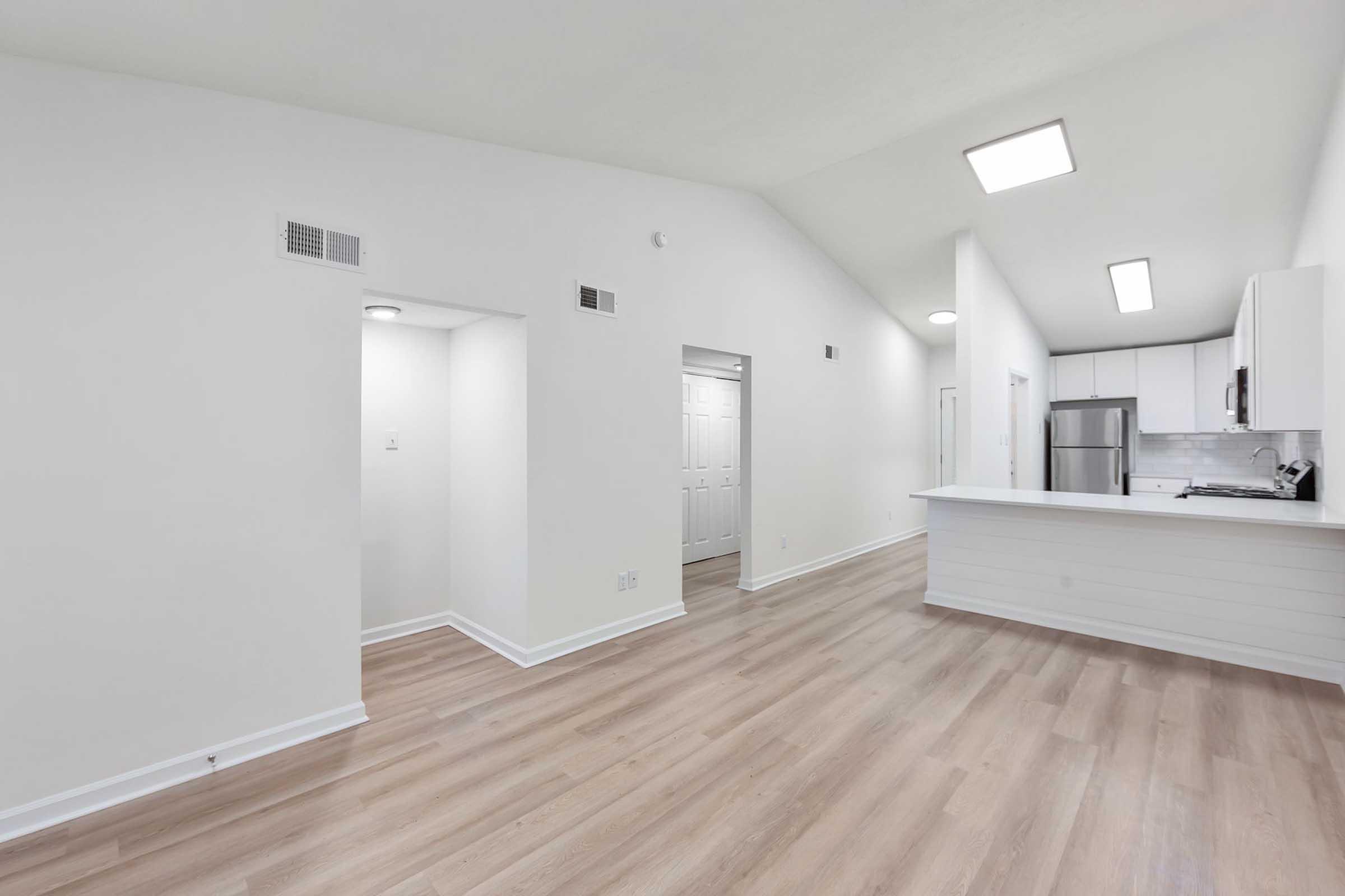 A spacious, modern interior of an apartment featuring white walls, light wood flooring, and high ceilings. The kitchen is open to the living area, showcasing a sleek design with stainless steel appliances. Doors lead to different rooms, emphasizing the airy layout and contemporary finishes.