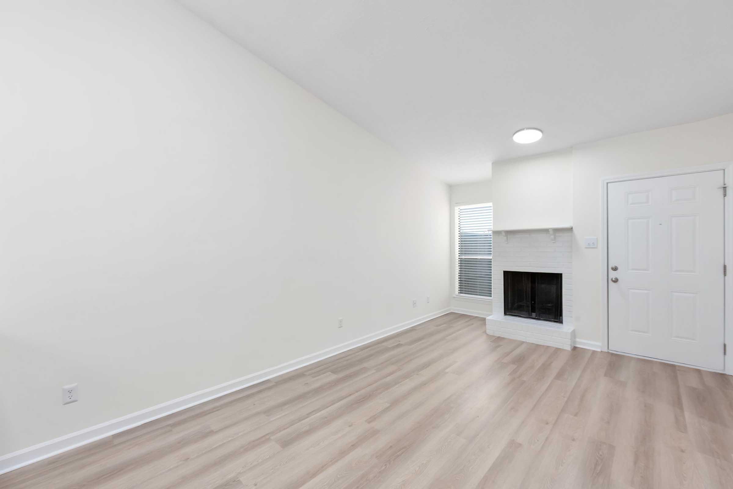 Empty living room with light-colored walls, hardwood flooring, and a white fireplace. A window with blinds allows natural light and the front door is visible on the right side of the image. The space appears bright and uncluttered, ready for furniture and decoration.