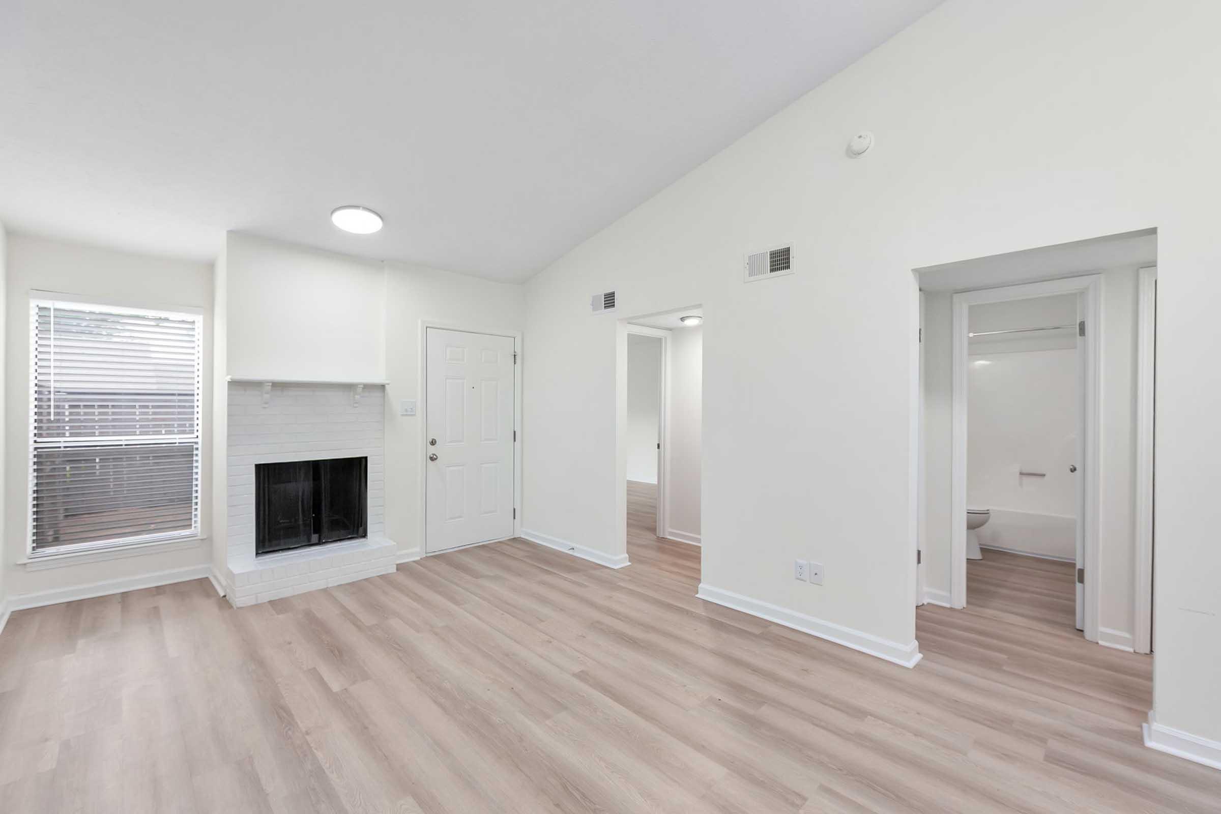 Spacious living room featuring light-colored walls, a white brick fireplace, and large windows with natural light. The room has vinyl flooring and leads to a hallway with doors to a bathroom and other rooms.