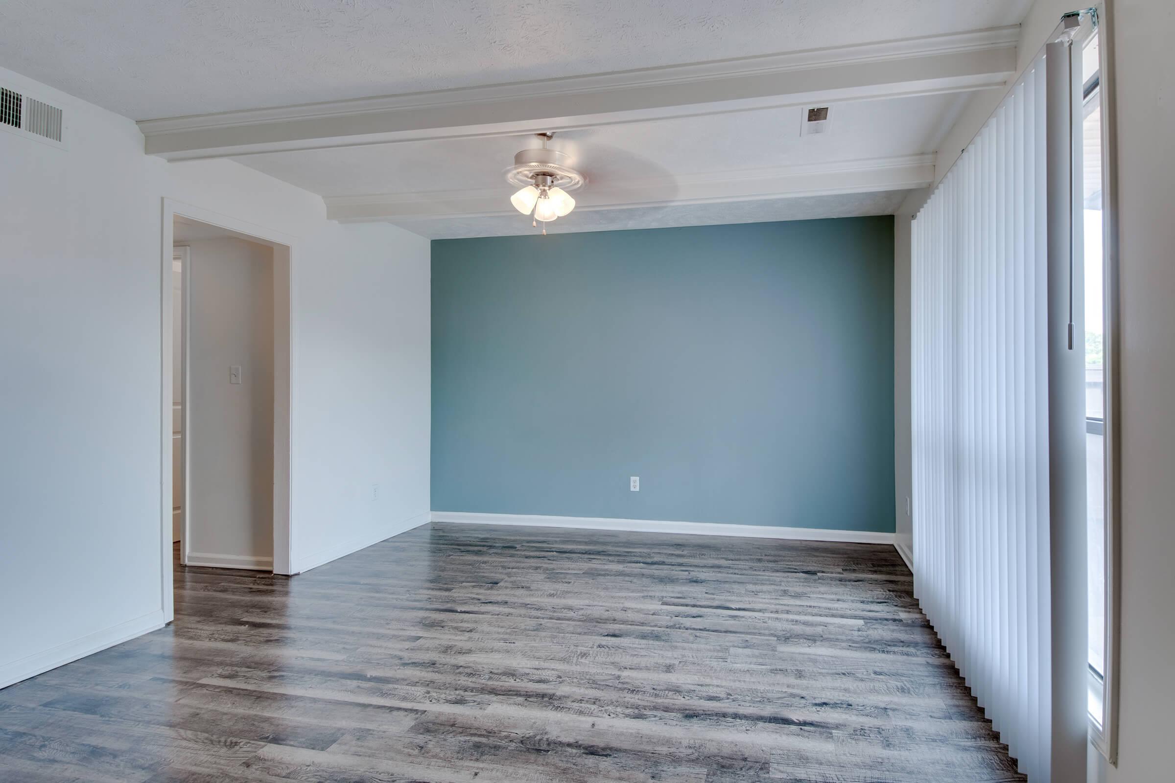 A spacious, well-lit room featuring a light blue accent wall and a ceiling fan. The flooring is dark wood, and large windows allow natural light. The room has an open layout, with transitions to other spaces visible through doorways.