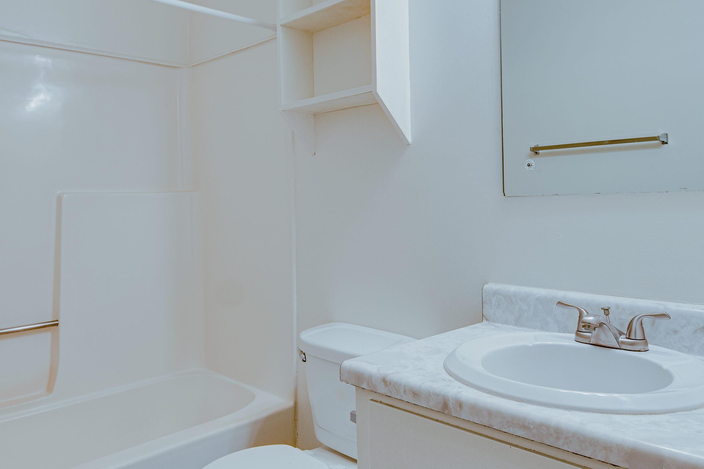 A clean, bright bathroom featuring a white bathtub and shower combination, a white toilet, and a sink with a marble countertop. A small shelf is mounted on the wall beside the mirror, and the overall color scheme is neutral, creating a spacious and tidy appearance.