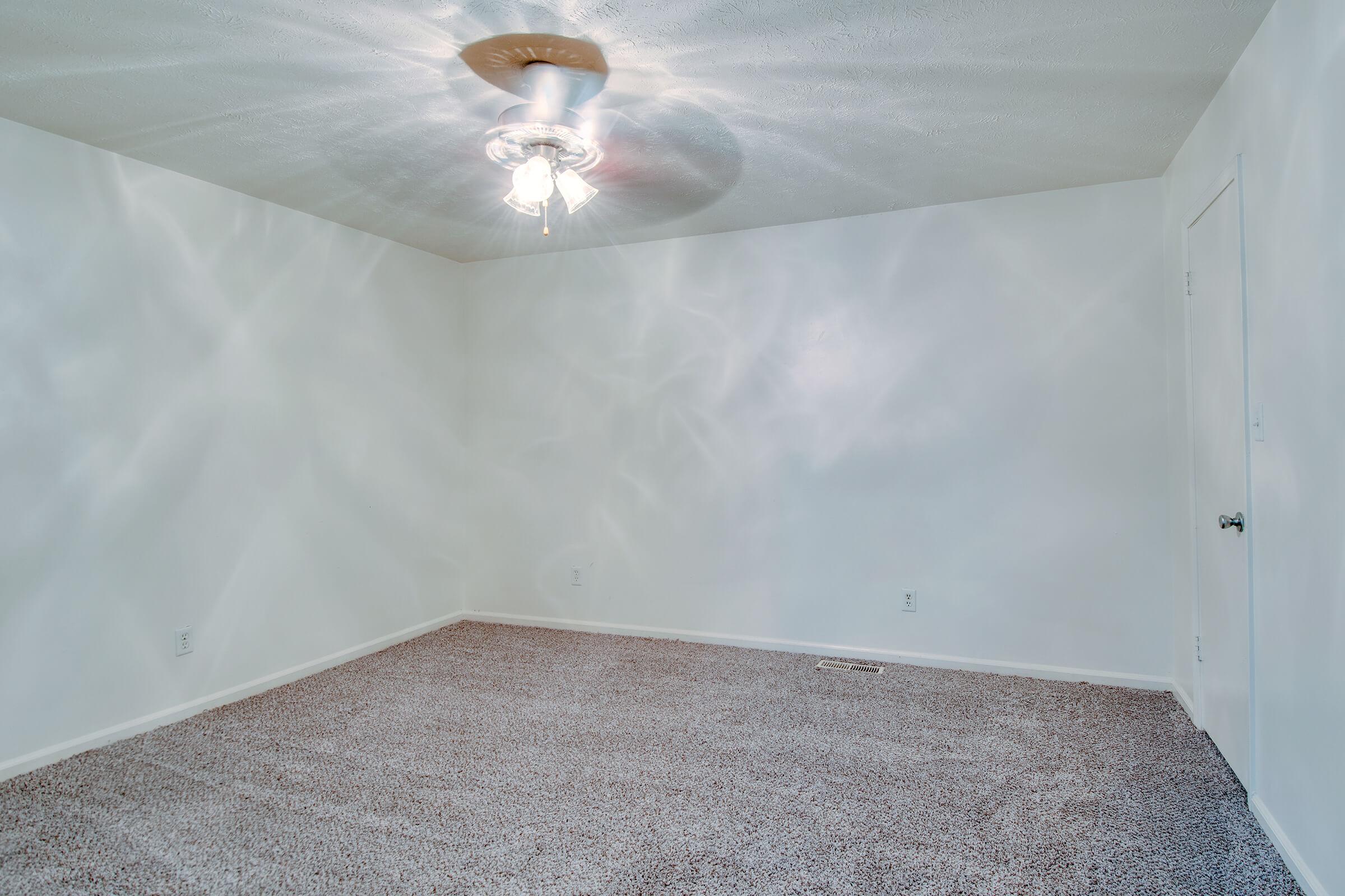 A spacious, empty room with freshly painted white walls and a light-colored carpet. A ceiling fan is installed in the center, casting soft light patterns on the walls. The room features a simple, uncluttered design and a closed door visible on the right.