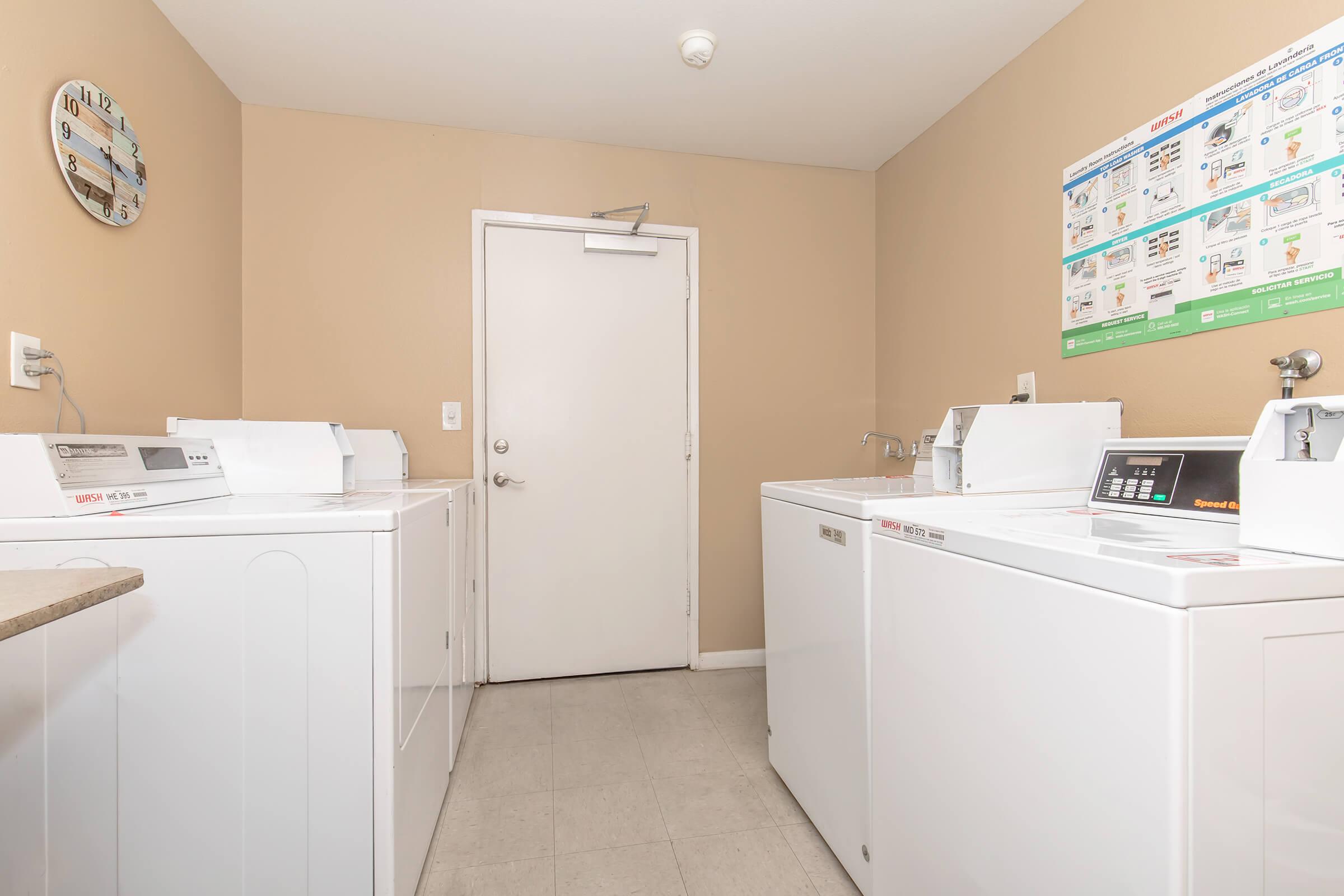 A clean laundry room with several white washing machines and dryers arranged against light-colored walls. A clock is mounted on the wall, and a door leads out of the room. Laundry instructions and information posters are displayed above the machines. The floor is tiled and the space is well-lit.