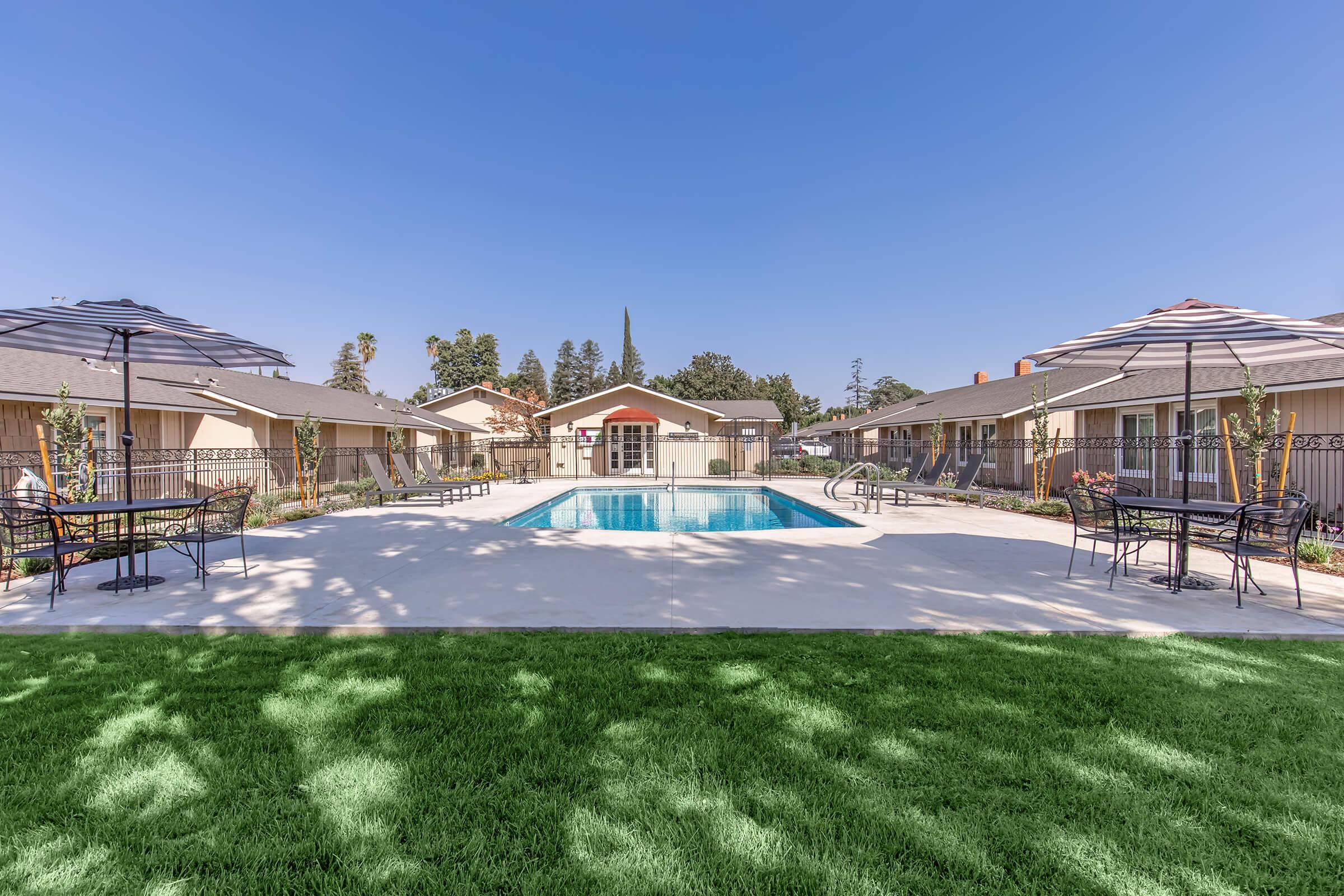 A clear blue sky above a pool area surrounded by landscaped gardens. The pool is rectangular and surrounded by lounge chairs and umbrellas. Nearby, there are several buildings with a residential appearance, all set on a well-maintained green lawn.