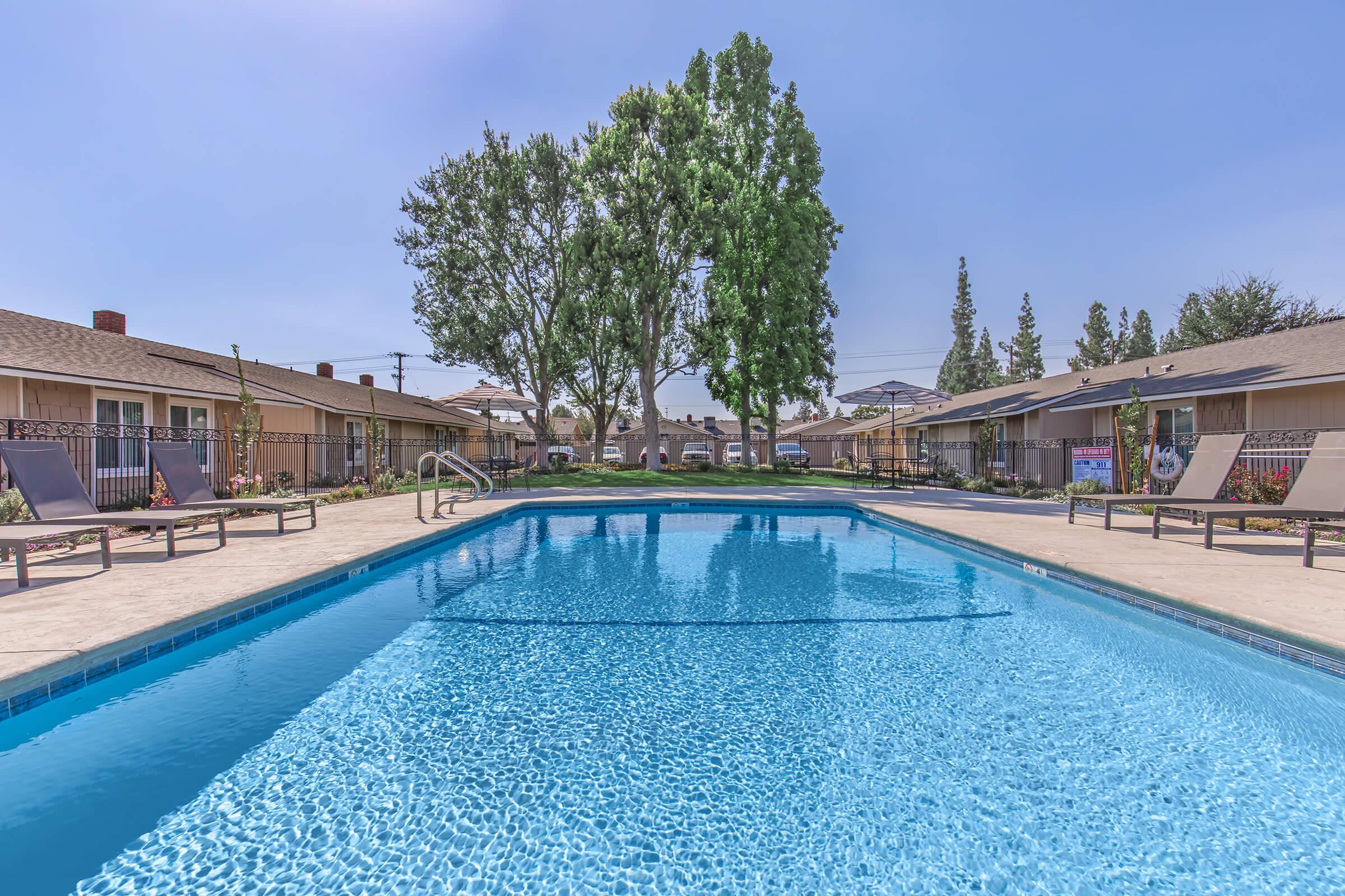 A clear swimming pool surrounded by lounge chairs and a grassy area. Tall trees provide shade in the background, with several residential buildings visible. The sky is bright blue, indicating a sunny day.