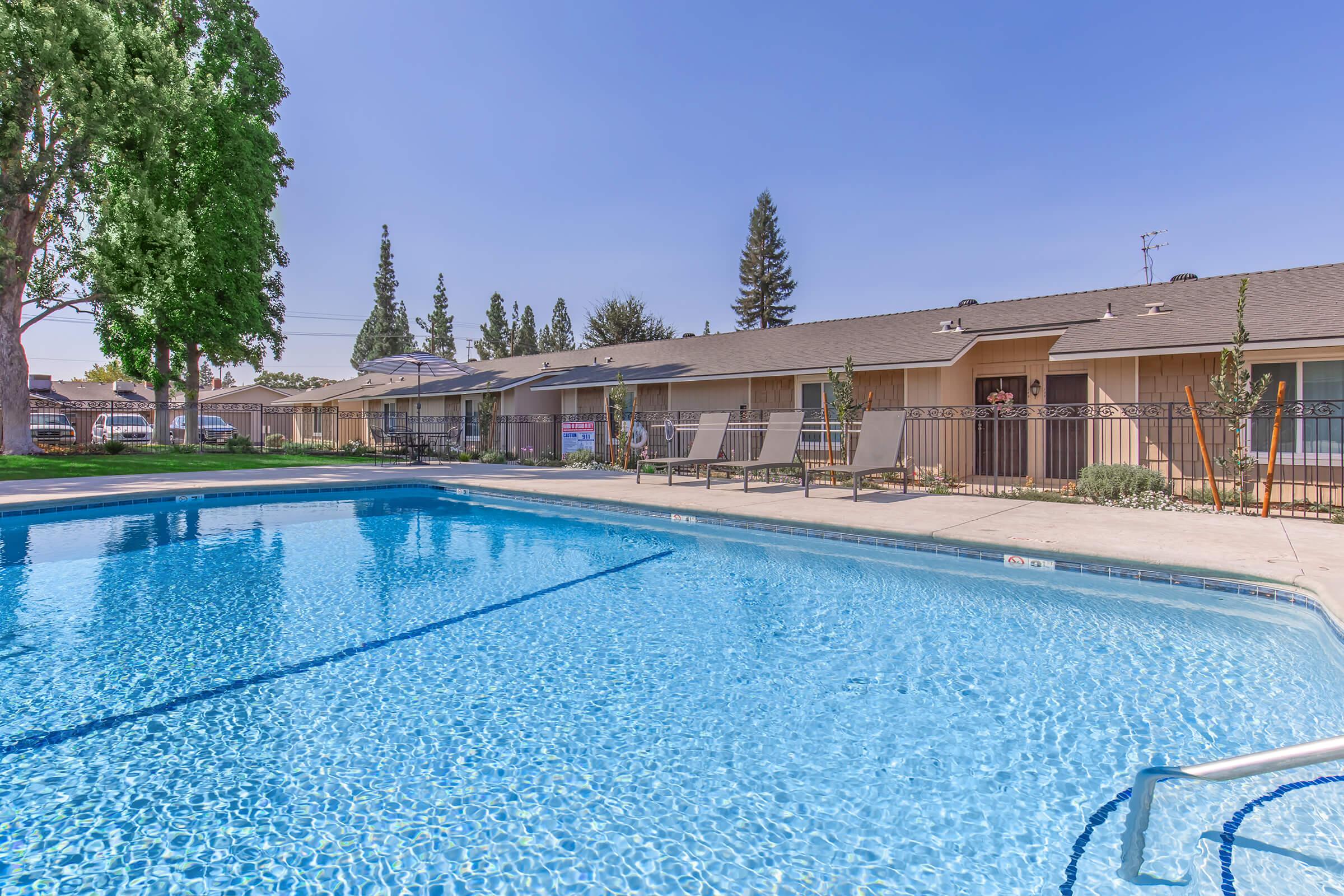 A swimming pool with clear blue water surrounded by a fence. Lounge chairs are positioned nearby on a sunlit day. In the background, there are residential buildings with a grassy area and trees, creating a relaxing atmosphere. The sky is clear and blue.