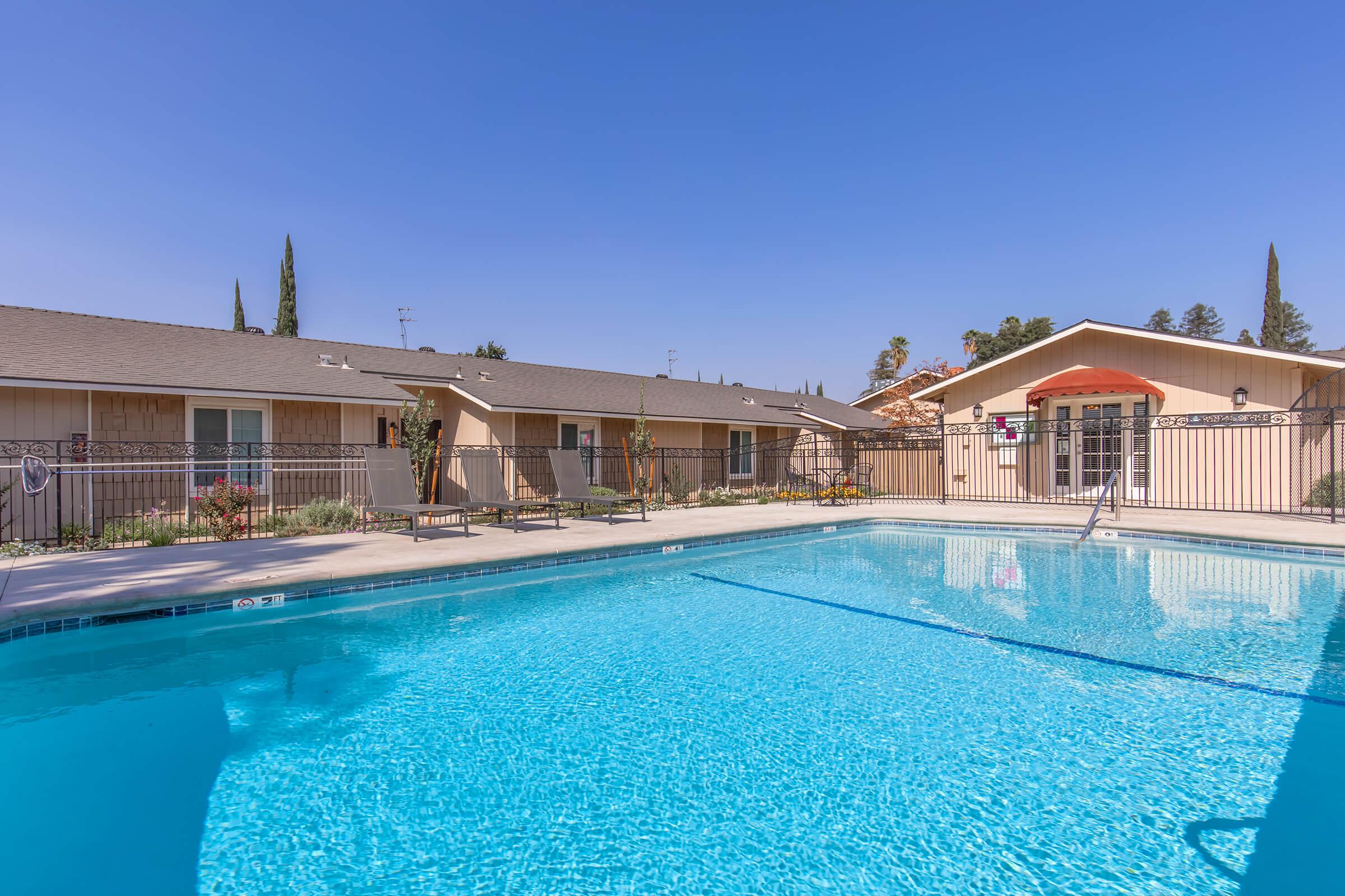 A bright blue swimming pool surrounded by lounge chairs and a fence, with a nearby building featuring a beige exterior and green trees in the background under a clear blue sky. The area is well-lit and inviting, ideal for relaxation and leisure activities.