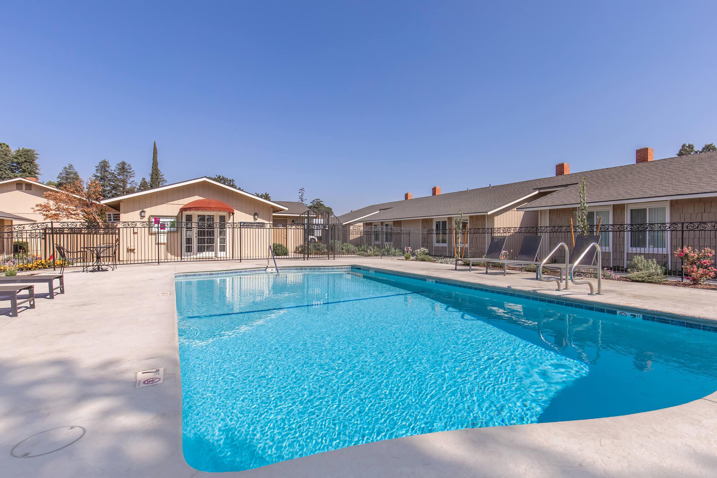 A clear blue swimming pool surrounded by a concrete deck, located in a residential area with beige buildings in the background. There are lounge chairs arranged nearby and landscaping elements like shrubs and small trees. The sky is bright blue with no visible clouds.