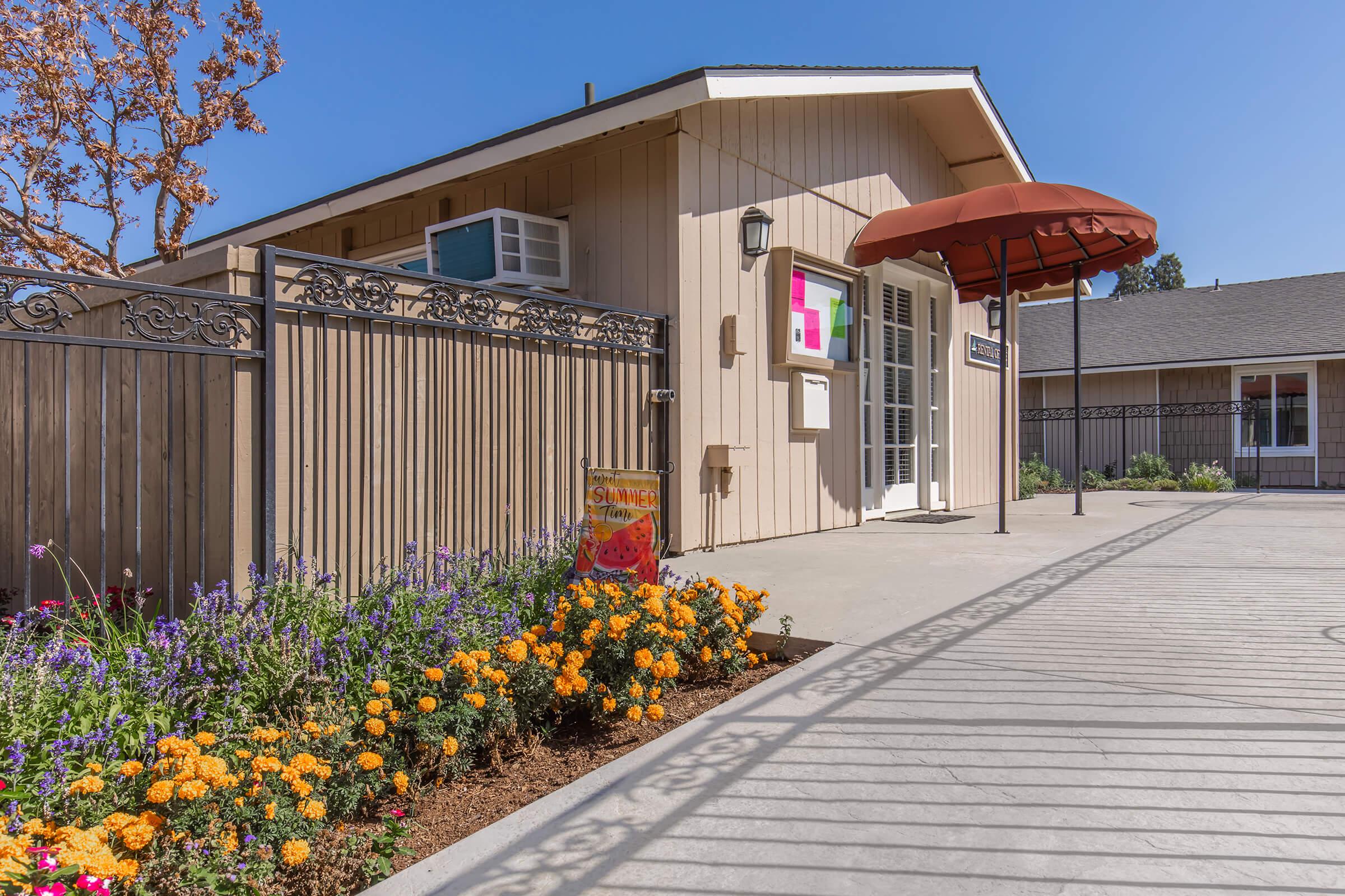 A tan-colored building with a red awning over the entrance. Colorful flowers in bloom are planted along the walkway, with orange marigolds prominently featured. The area has a well-maintained appearance, surrounded by a decorative fence, and blue skies in the background.