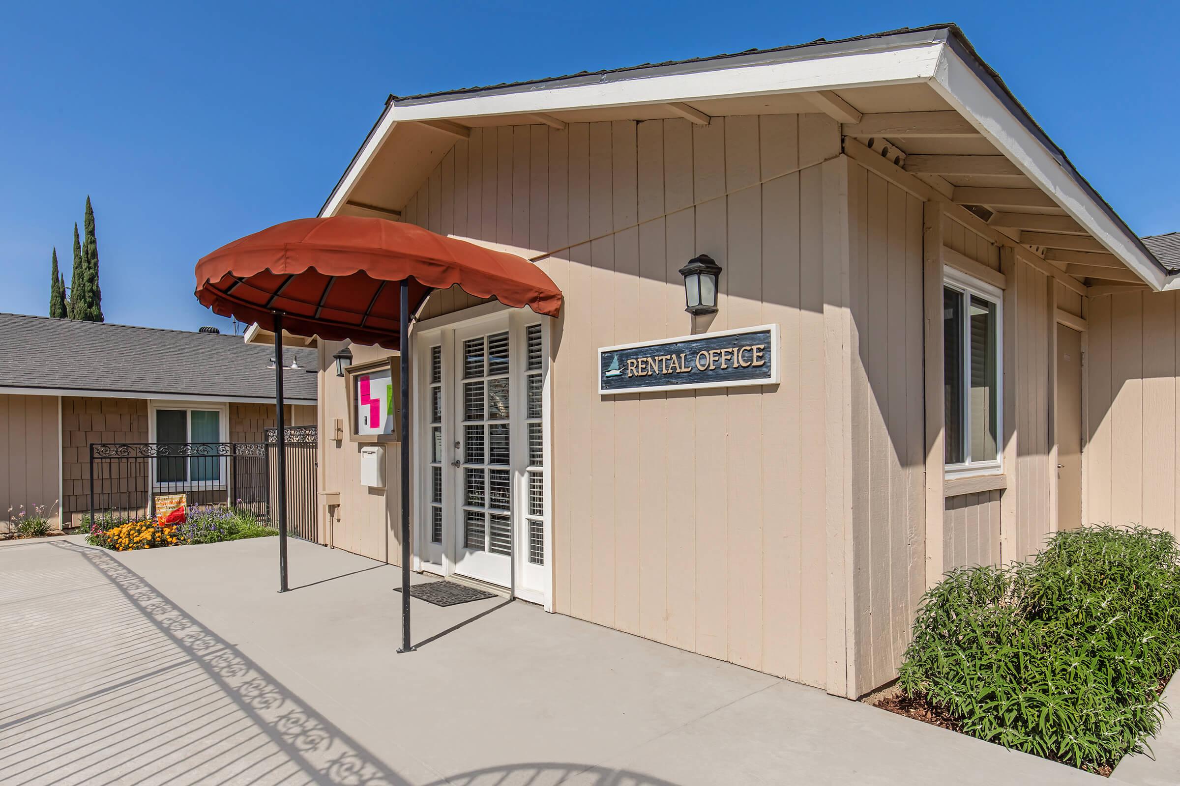 A rental office building with a tan exterior and a red awning. The entrance features double doors with glass panels, a sign reading "RENTAL OFFICE," and a light fixture beside the door. There are landscaped flower beds and a walkway leading to the entrance under a clear blue sky.