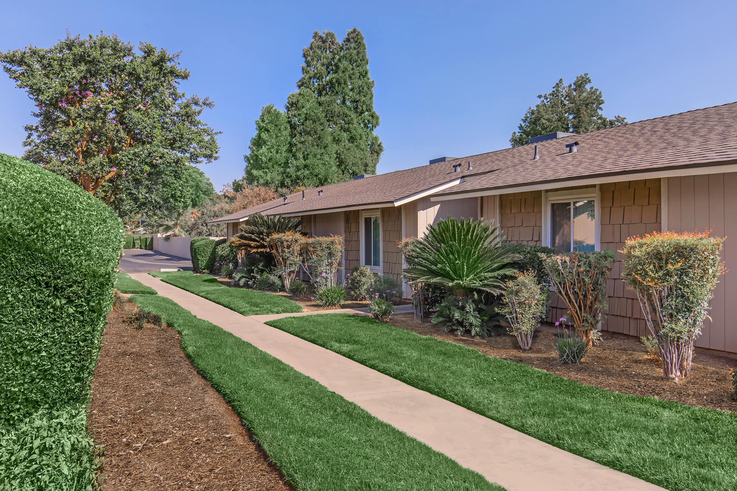 A landscaped pathway leading to a row of single-story residential units. The area features manicured grass, decorative shrubs, and palm trees, providing a serene and inviting atmosphere. The sky is clear, contributing to the pleasant setting of the neighborhood.