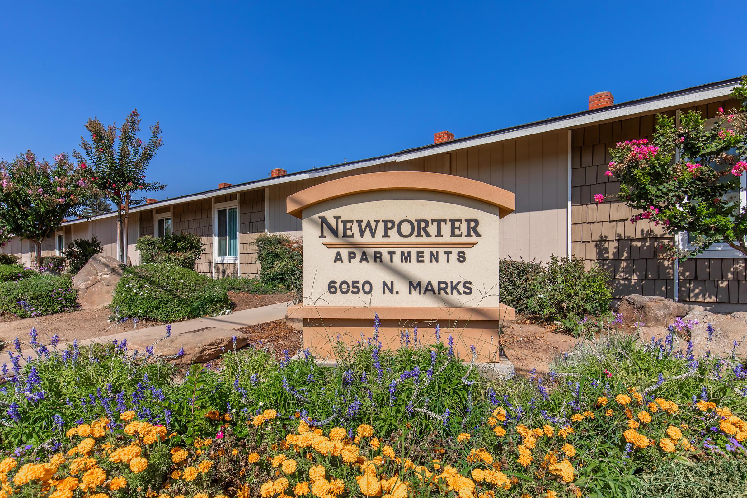 Sign for Newporter Apartments at 6050 N. Marks, surrounded by colorful flowers and greenery. The building features a modern architectural design with a clear blue sky in the background.