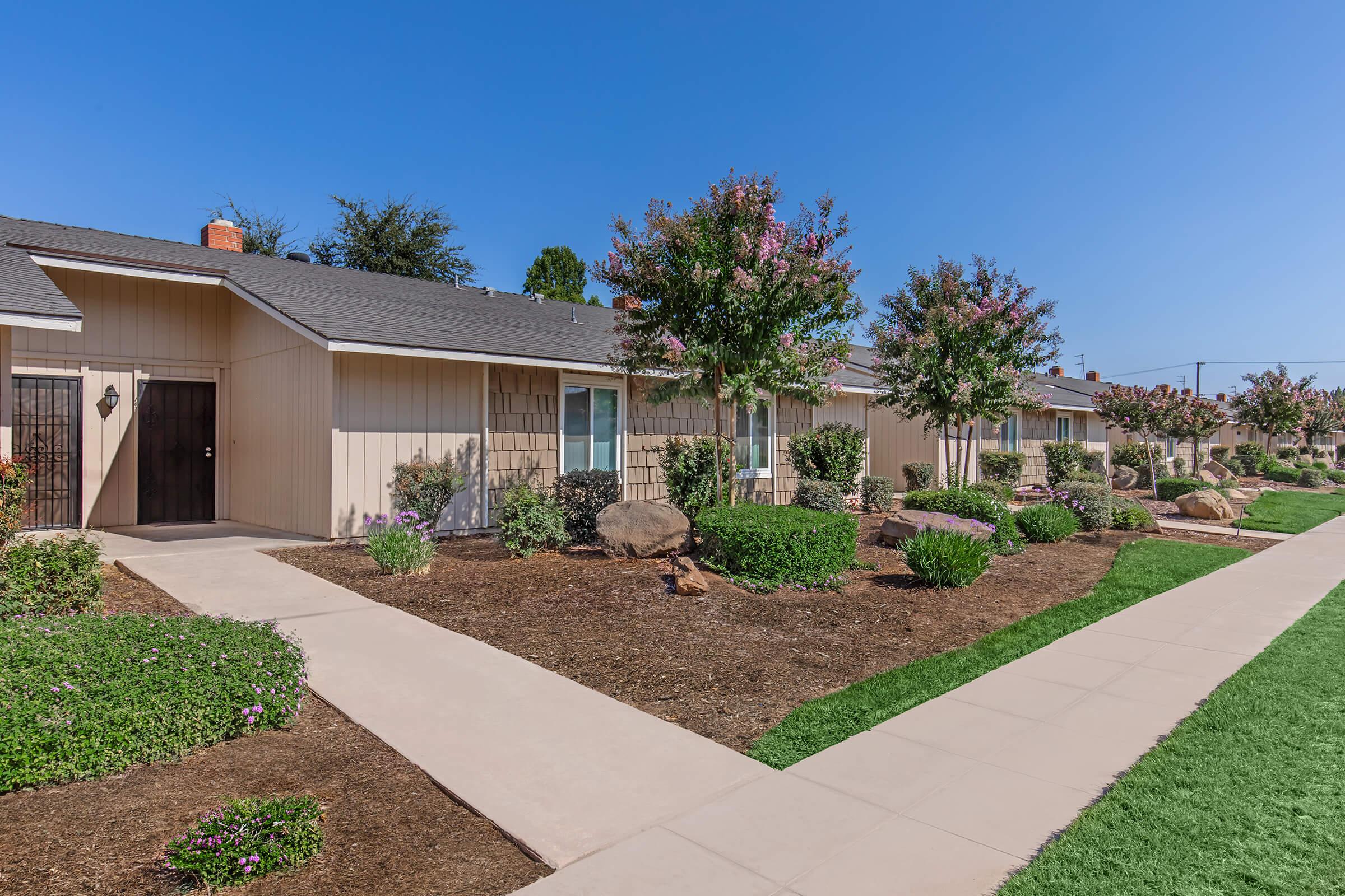 A row of single-story residential buildings with landscaped gardens. Each unit features a front door and window, surrounded by bushes and small trees. A wide concrete pathway runs alongside the buildings, leading through a manicured lawn and flower beds under clear blue sky.
