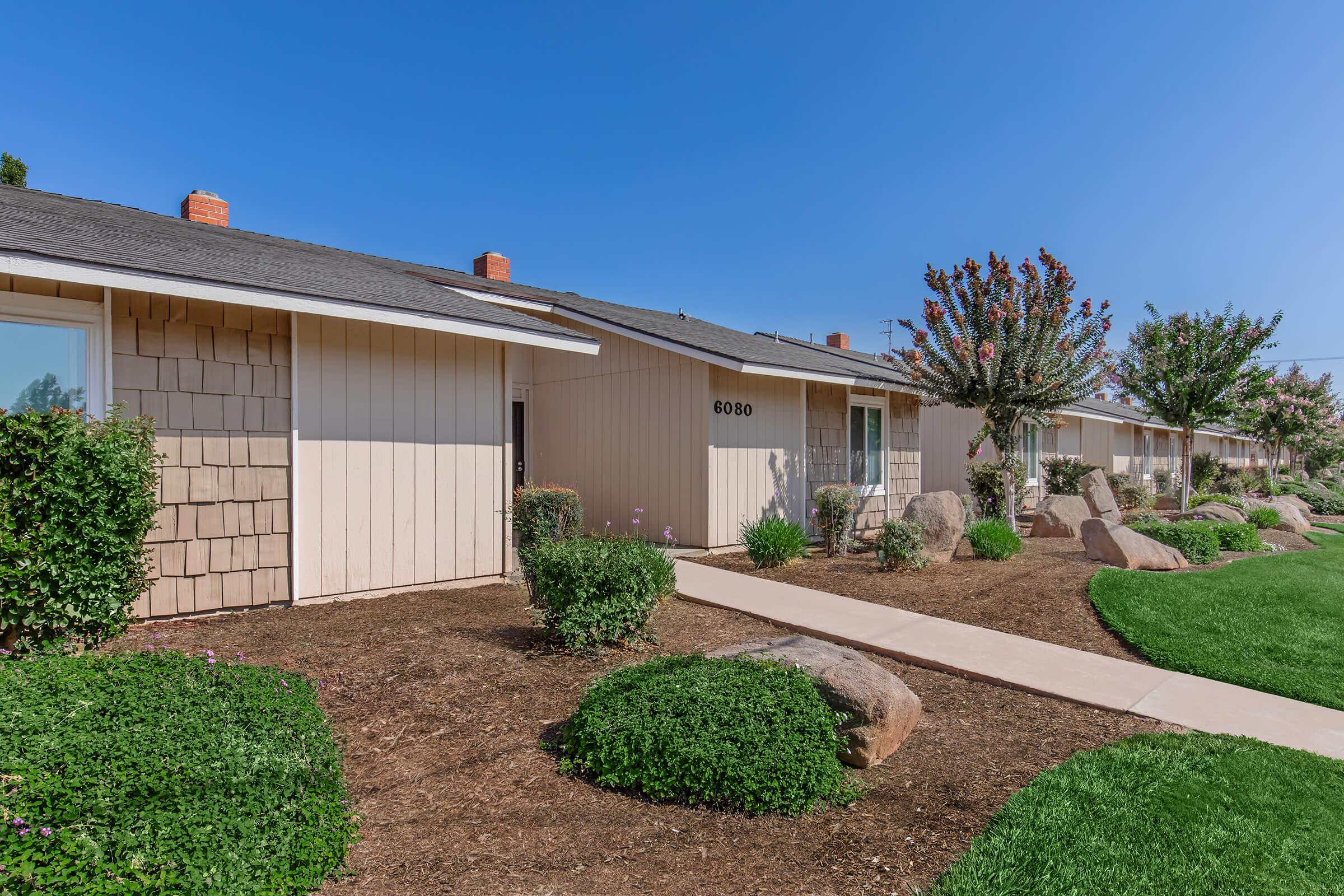 A row of modern single-story houses with light-colored siding, neatly landscaped front yards, and manicured bushes. The clear blue sky is visible above, and there are a few trees and rocks in the landscaping, enhancing the serene outdoor atmosphere. Pathways lead to the entrances of the homes.
