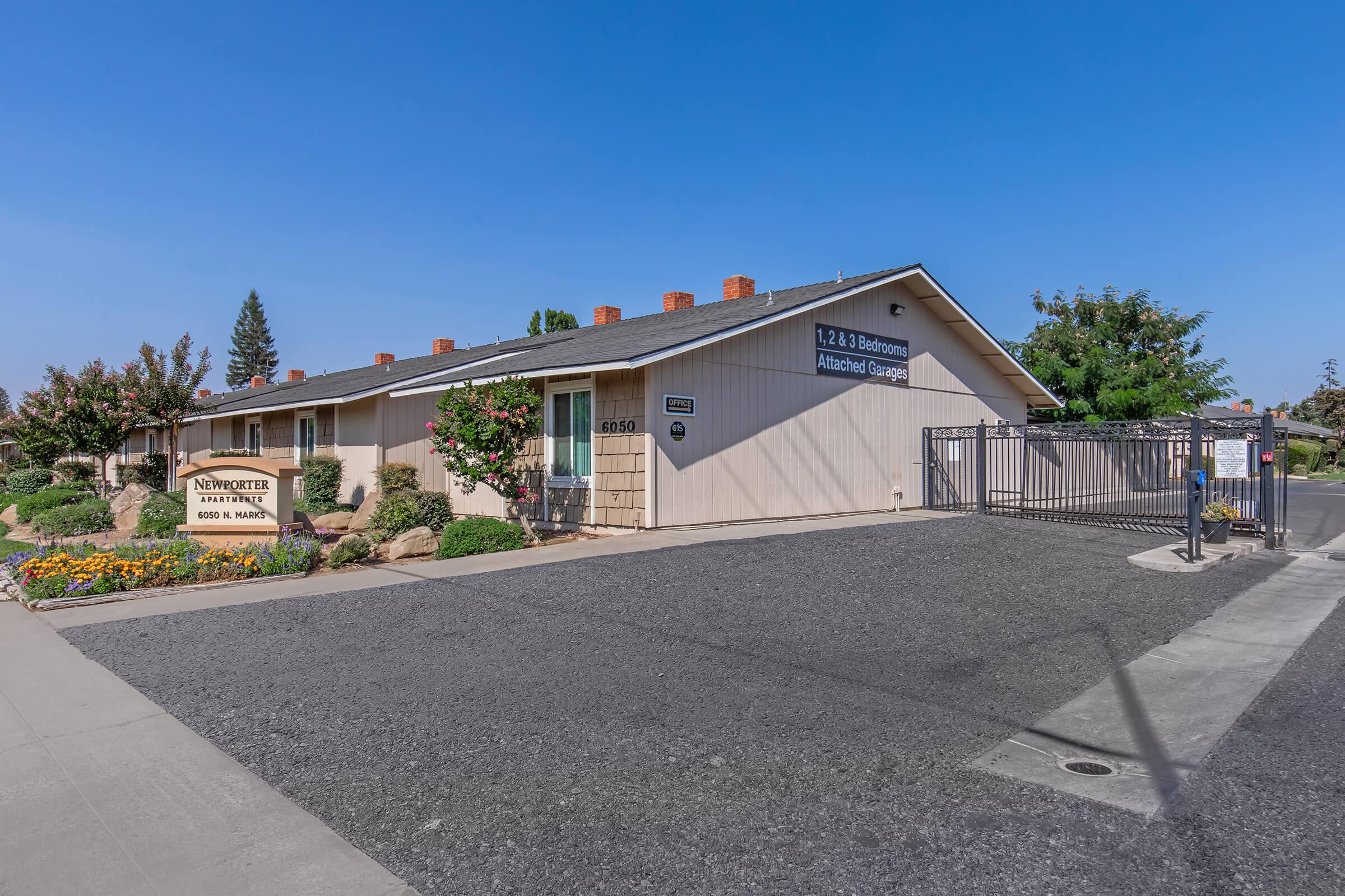 A single-story building with a tan exterior and a peaked roof, featuring a welcoming entrance with flowers and shrubs. A sign indicates it's an "Independent Living Facility." The property has a gated entrance and is surrounded by landscaping and an asphalt parking area. Clear blue sky above.