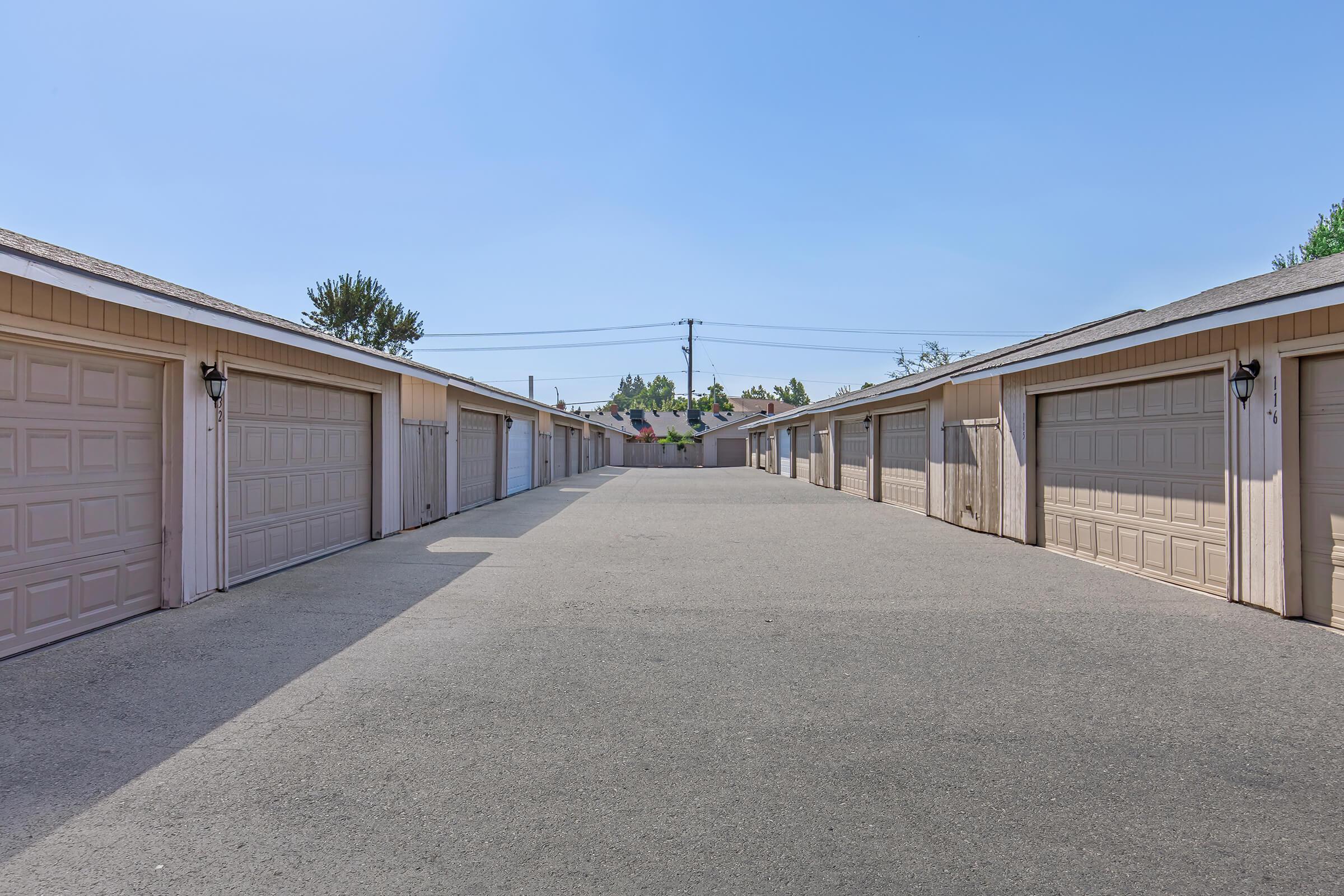 A wide-angle view of a row of garages with closed garage doors on either side of a paved driveway. The sky is clear and blue, and there are trees in the background. The scene conveys a quiet and spacious atmosphere, typical of a residential area.