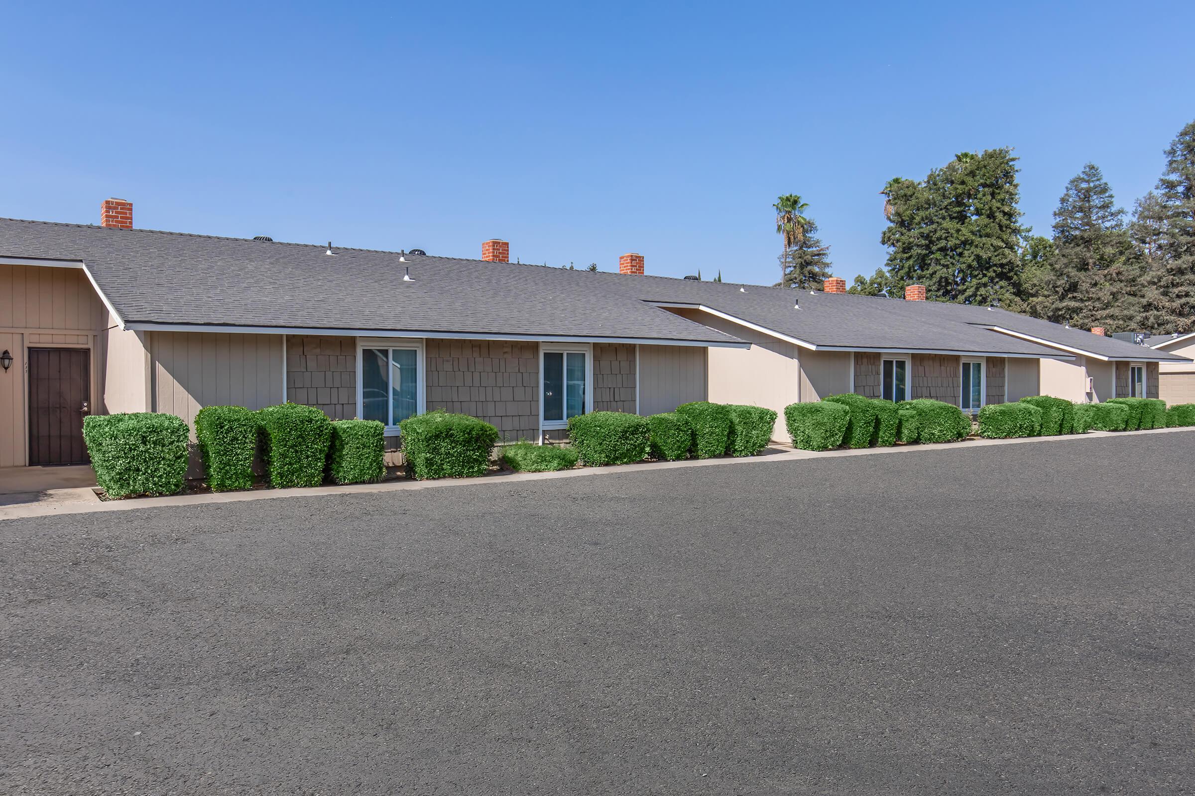 Single-story motel-style building with several rooms, featuring light-colored siding and dark shingle roofs. Neatly trimmed green shrubs line the pathway in front, and a clear blue sky is visible above. The parking area is paved and empty, providing a clean and orderly appearance.