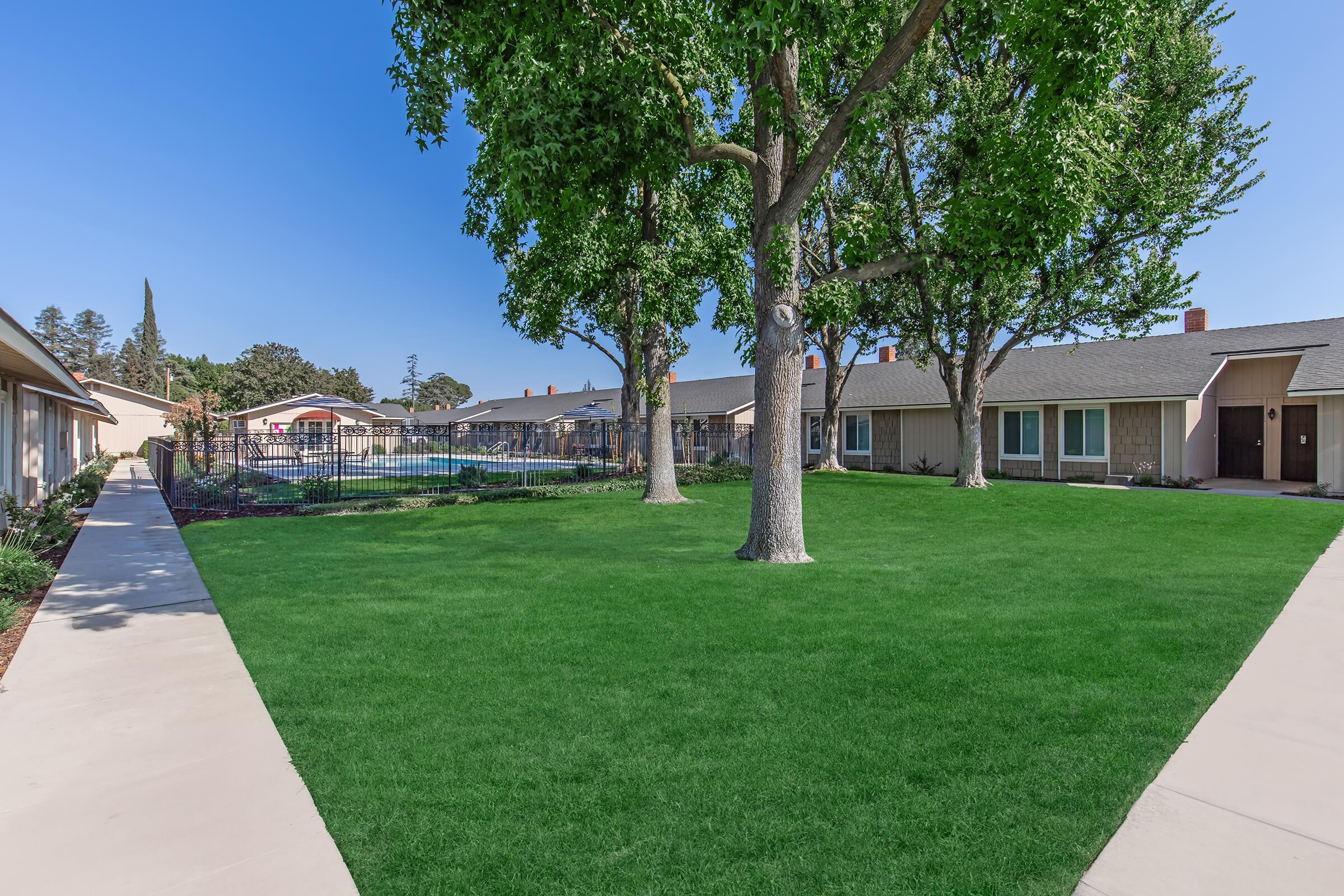 A well-maintained courtyard featuring a green lawn surrounded by pathways. In the background, there is a swimming pool with a fence, flanked by single-story buildings. Large trees provide shade, and the sky is clear, indicating a sunny day.