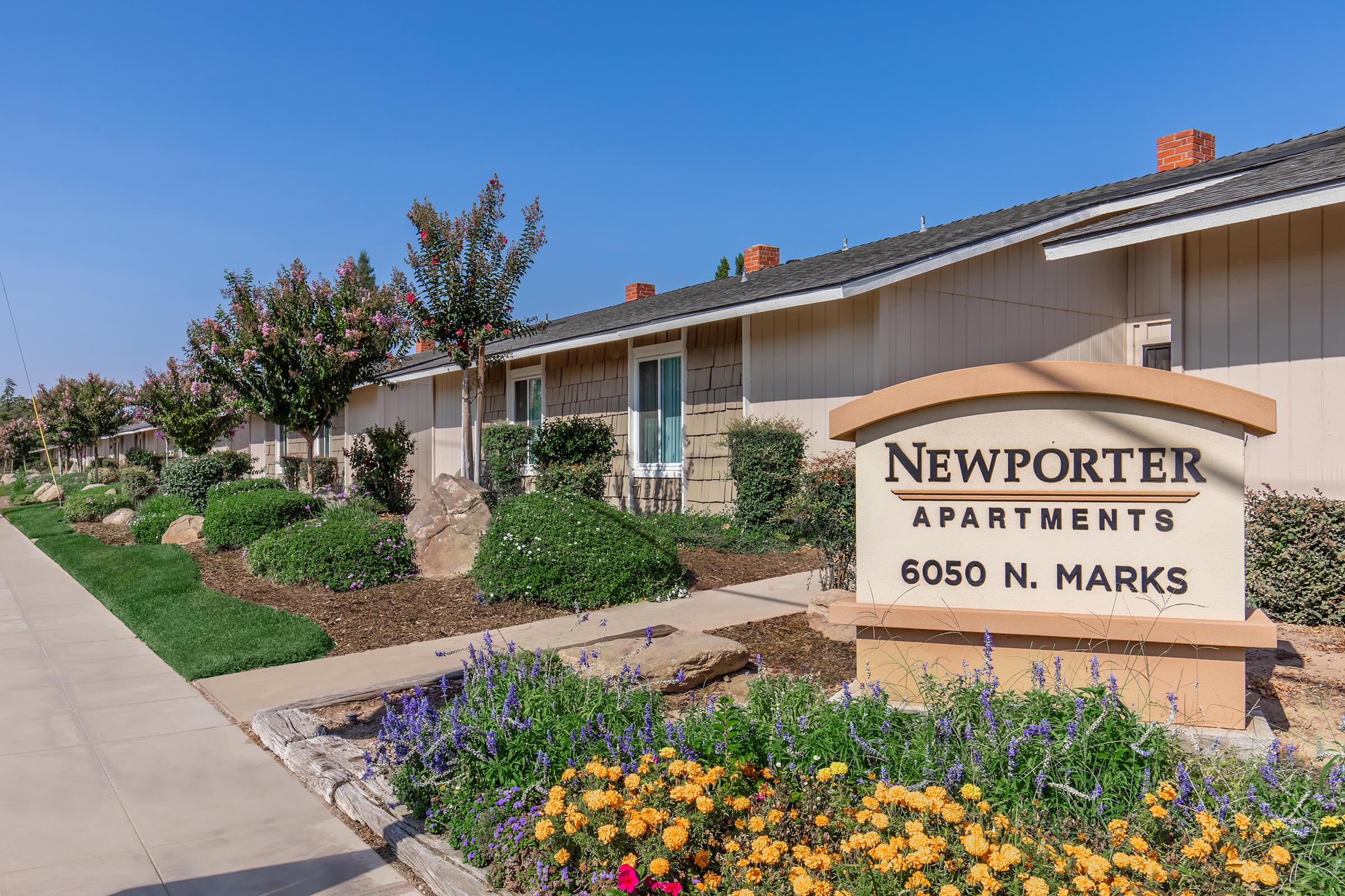 Exterior view of the Newporter Apartments, featuring a sign displaying the name and address. The building has landscaped gardens with colorful flowers, shrubs, and trees. A clear blue sky is visible above, and a sidewalk runs along the property.