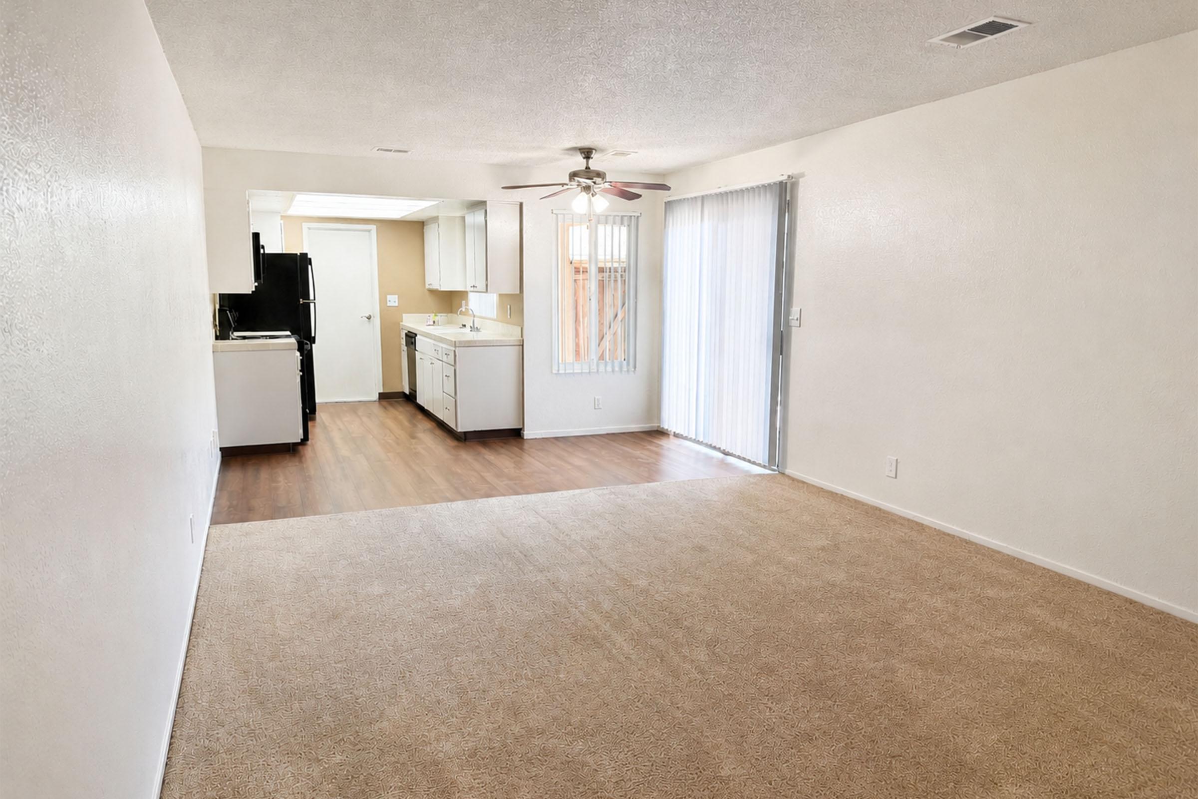 Spacious living area with light-colored walls and carpet. To one side, there’s a kitchen featuring white cabinets and black appliances. A sliding glass door allows natural light to enter and provides access to an outdoor area. The overall atmosphere is bright and inviting, ideal for comfortable living.