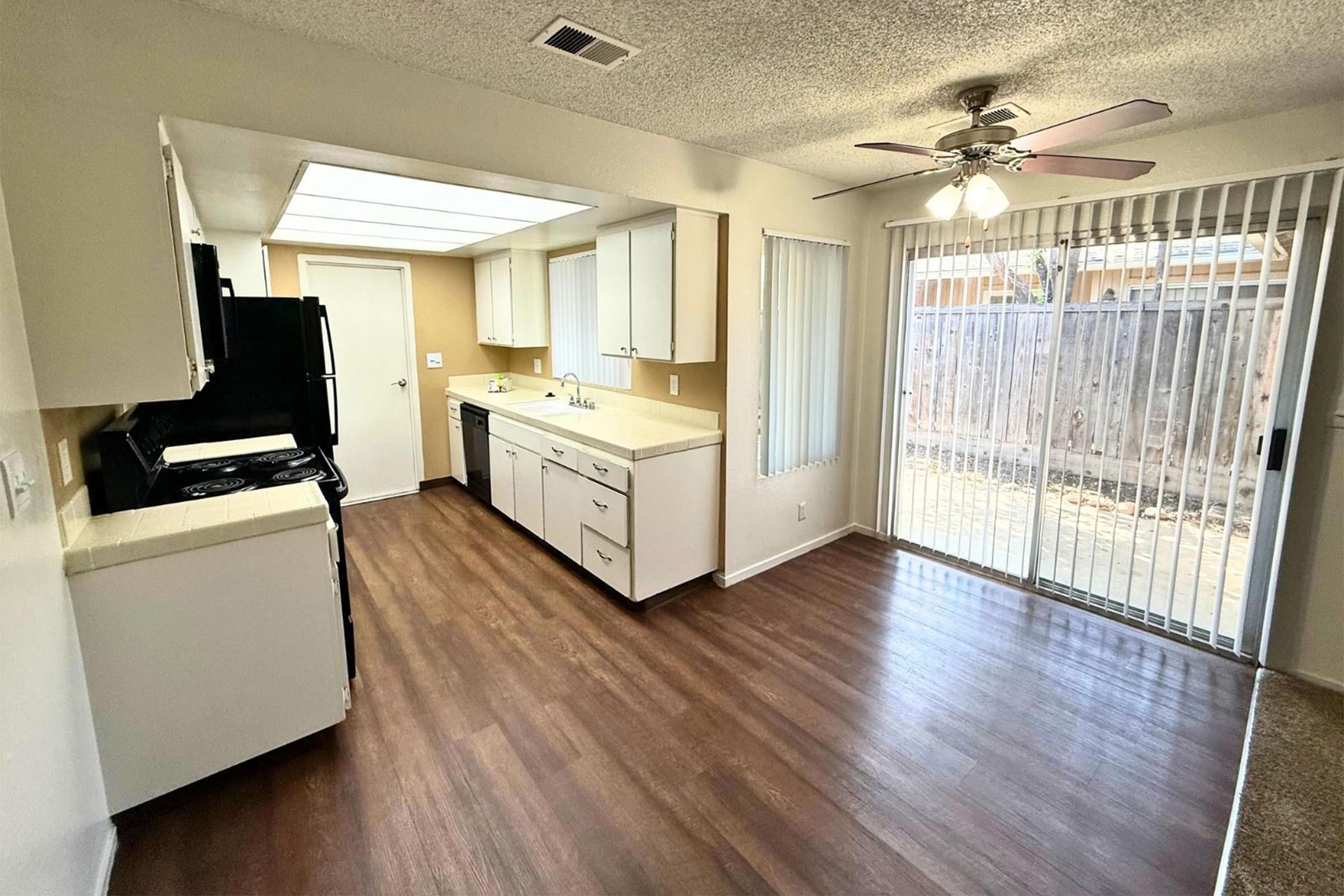 A spacious kitchen with white cabinets and a black stove and refrigerator. Natural light filters in through sliding glass doors leading to an outdoor area. The room features wooden flooring and a ceiling fan, creating a bright and airy atmosphere.
