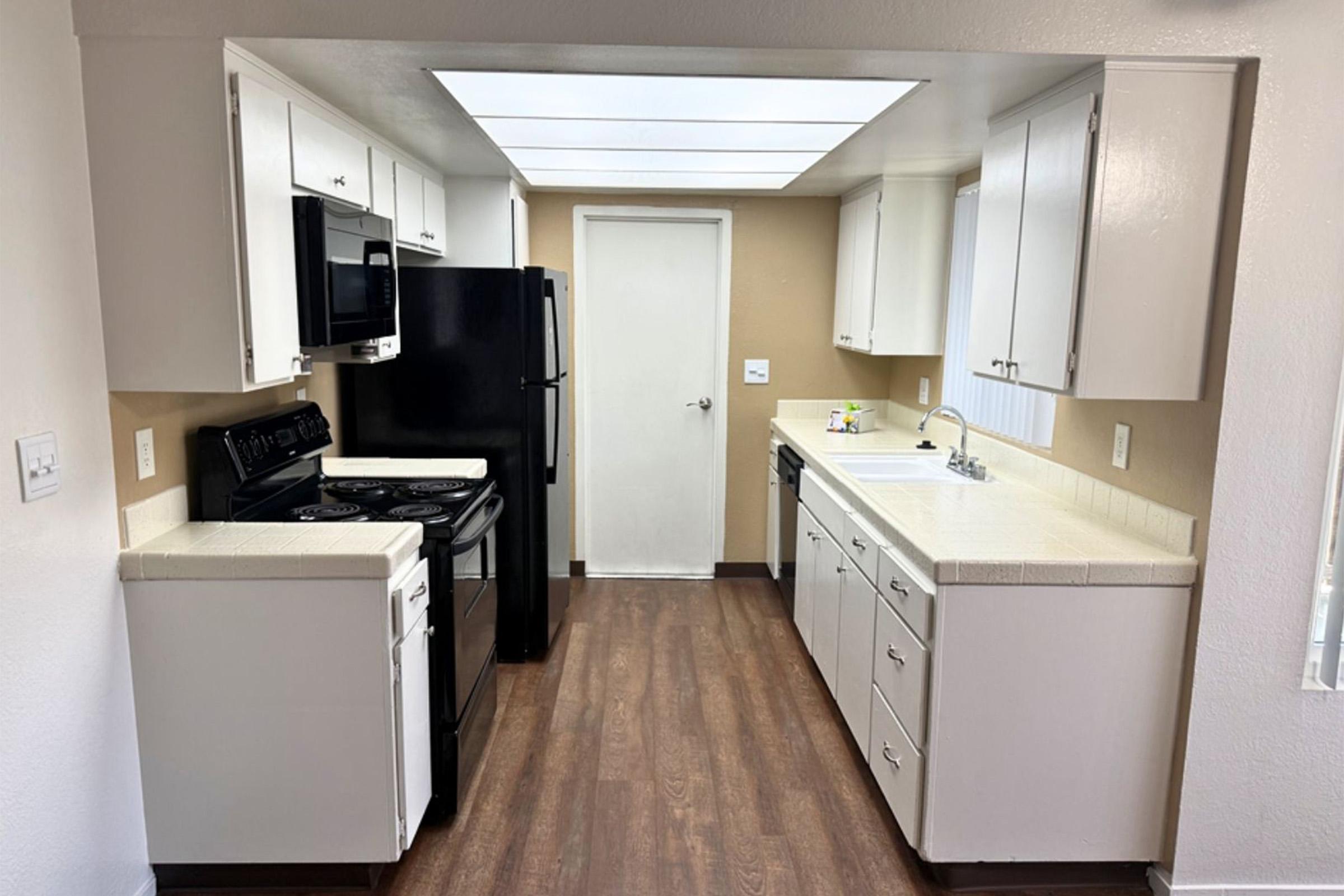 A modern kitchen featuring white cabinets, a black refrigerator, and a black oven with a stovetop. The countertops are light-colored, and there's a double sink under a kitchen window. The walls are painted in a warm beige, and the floor has a wood-like laminate. Accessible door leads to another room.