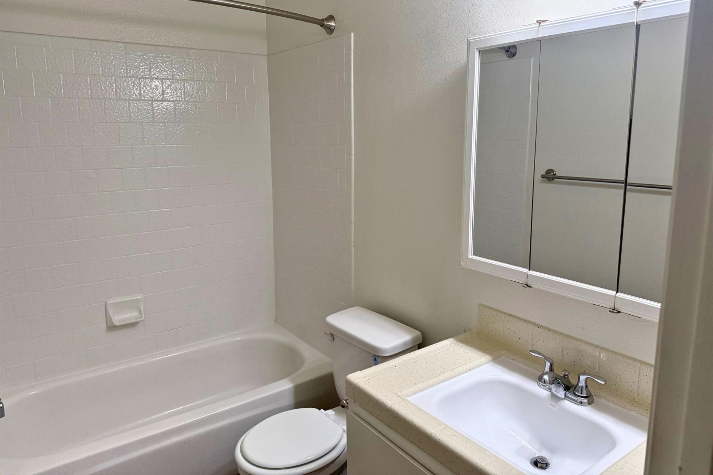 A clean bathroom featuring a white bathtub and shower combination, a toilet, and a sink with a light-colored countertop. The walls are tiled in a simple, white pattern, and a mirror is mounted above the sink. Natural light can be seen reflecting off the surfaces.