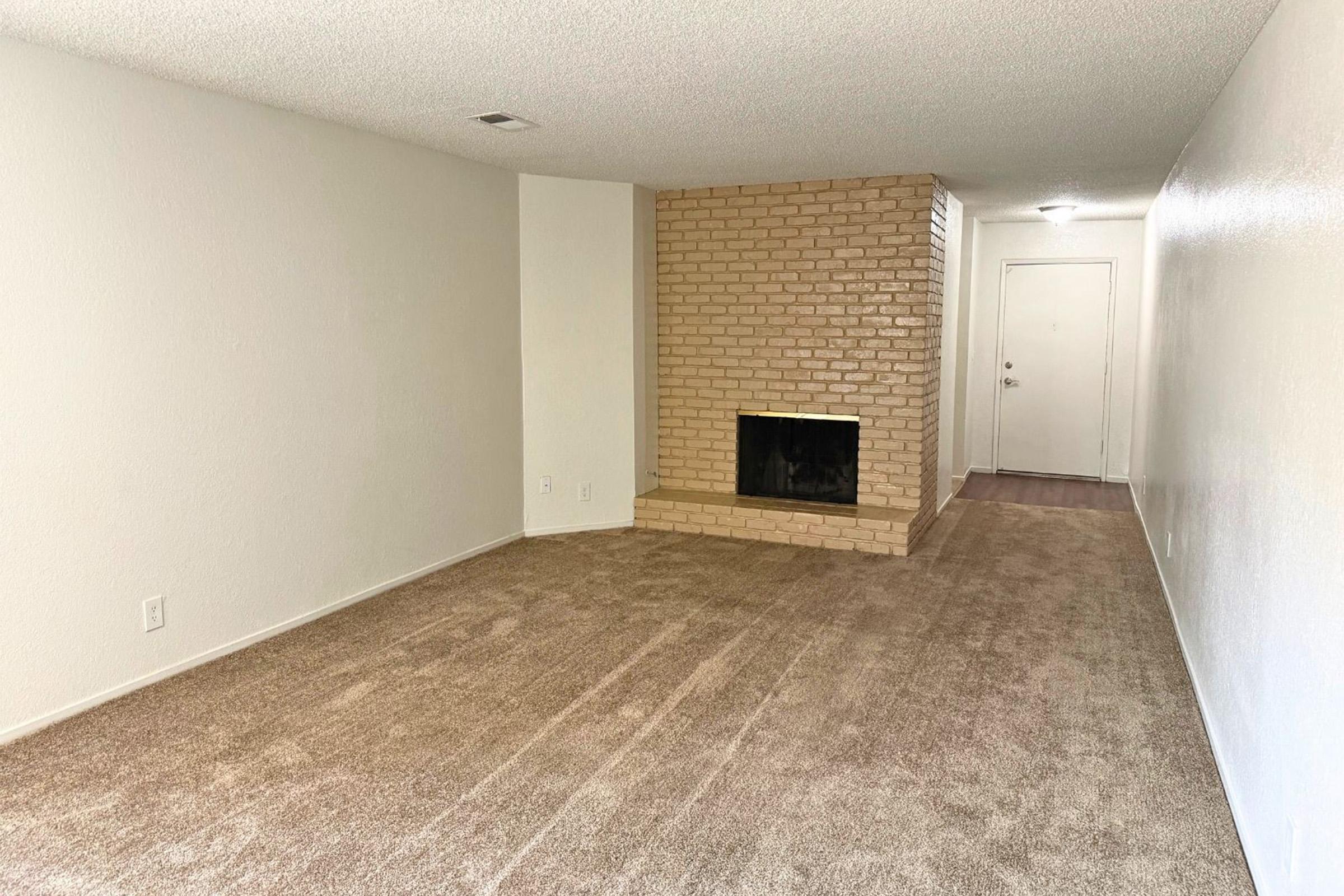 A spacious living room with beige carpeted flooring and white walls. A brick fireplace serves as a central feature against one wall. There's a door on the right side leading outside, and the room appears bright with natural light. The overall atmosphere is clean and inviting, suitable for various furnishings.